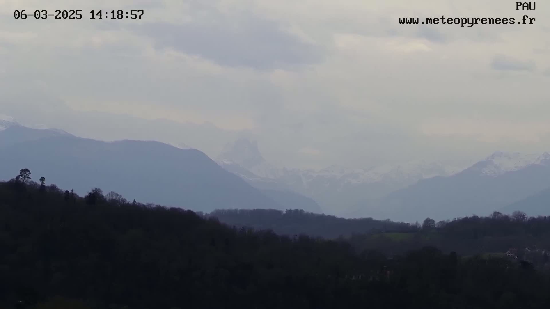 A hazy, overcast day reveals a view of distant, snow-capped mountains behind a foreground of dark, silhouetted hills and trees.