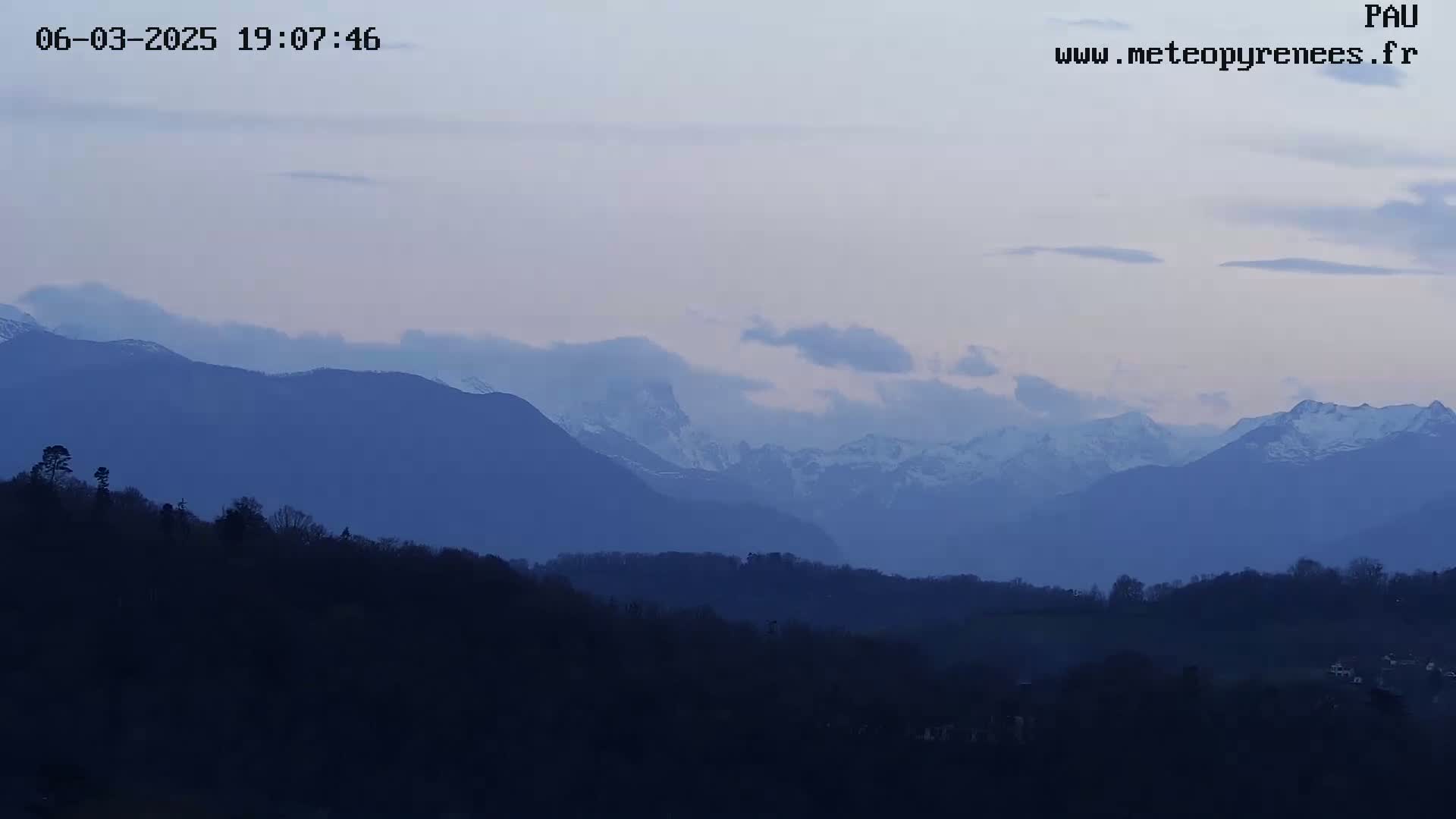 A view of snow-capped mountains in the distance, silhouetted against a pale twilight sky, with a foreground of dark, rolling hills.