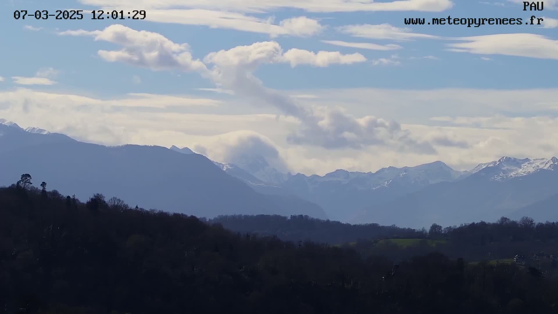A partly cloudy sky overlooks a dark, forested hillside in the foreground and snow-capped mountains in the distance.