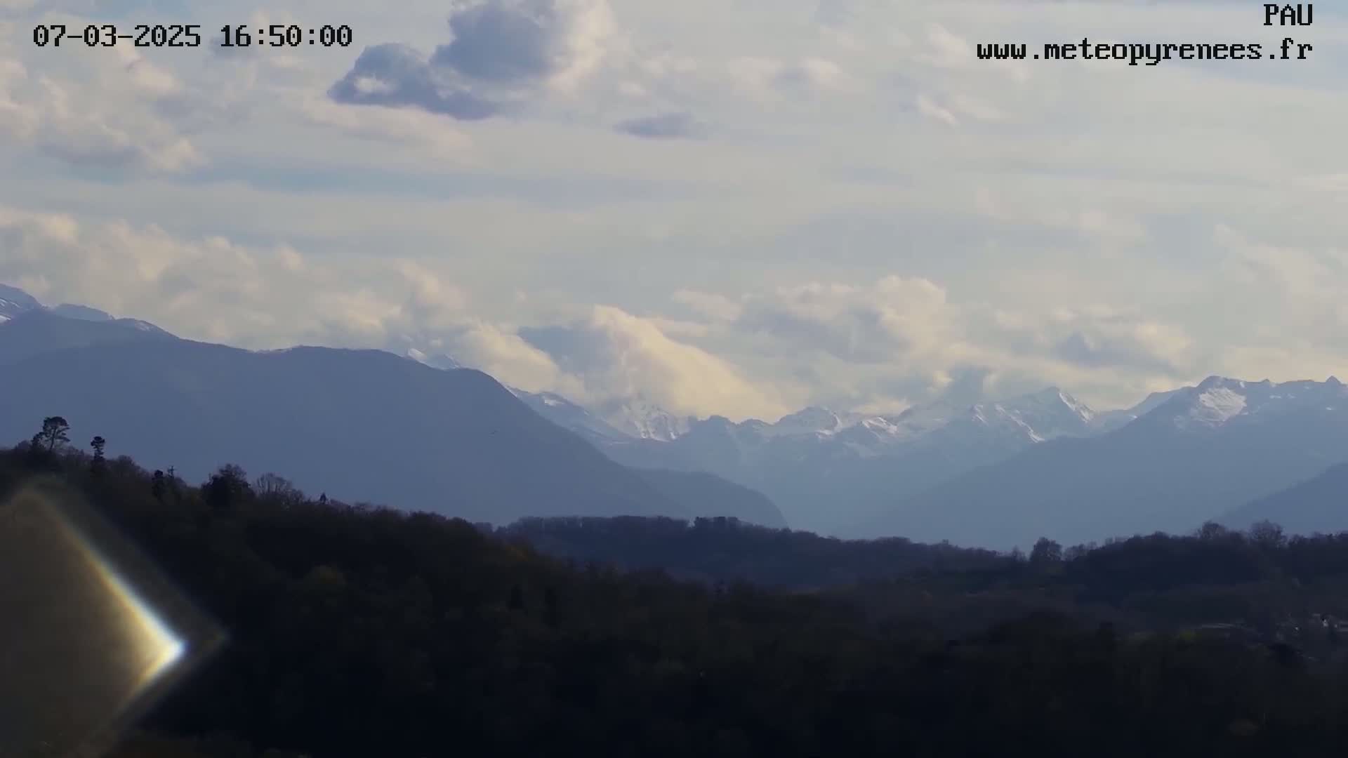 A partly cloudy sky overlooks a forest and distant, snow-capped mountains.