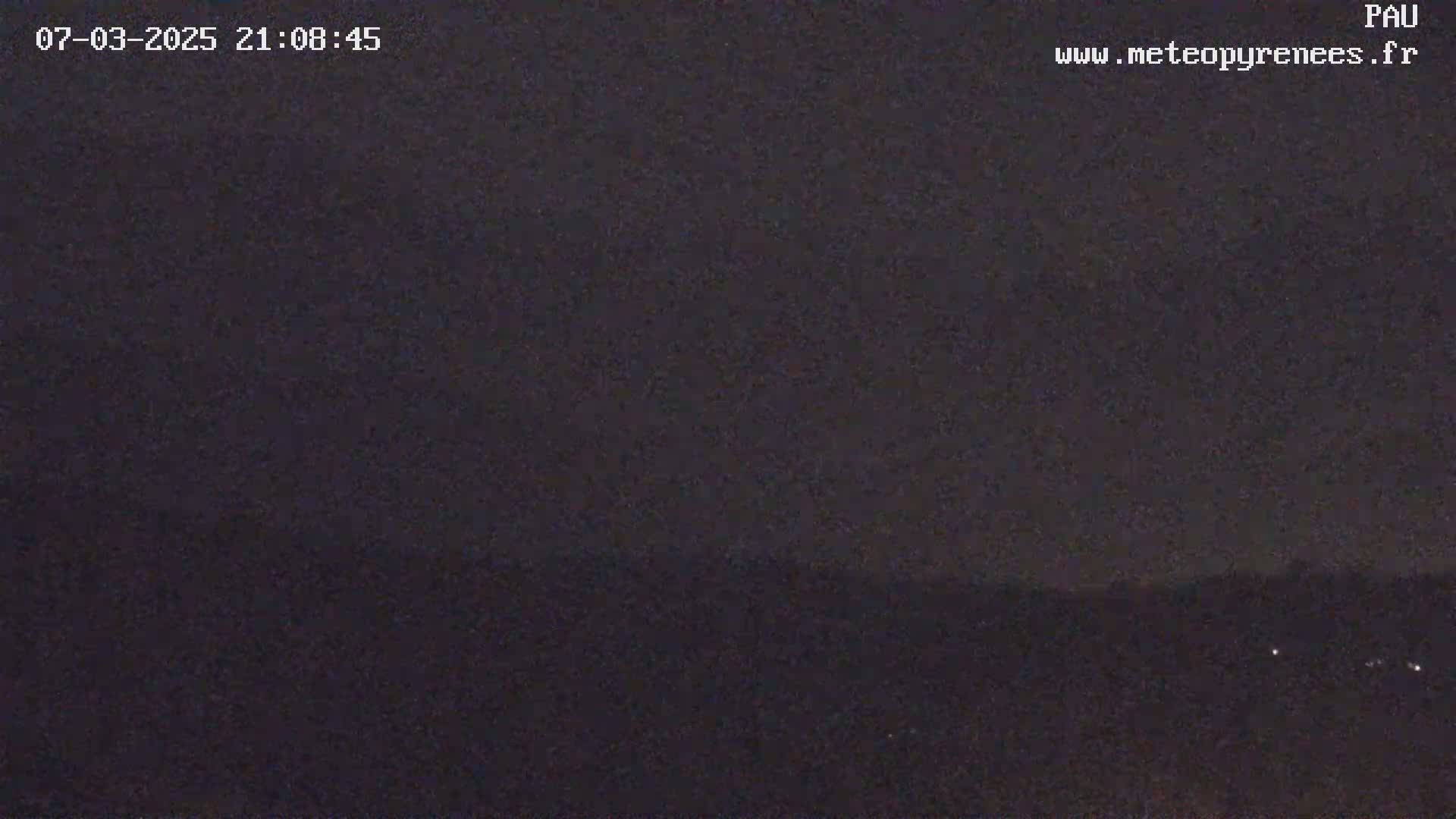 A nighttime long shot shows a dark landscape under a mostly clear, dark sky with a few faint lights visible in the distance.