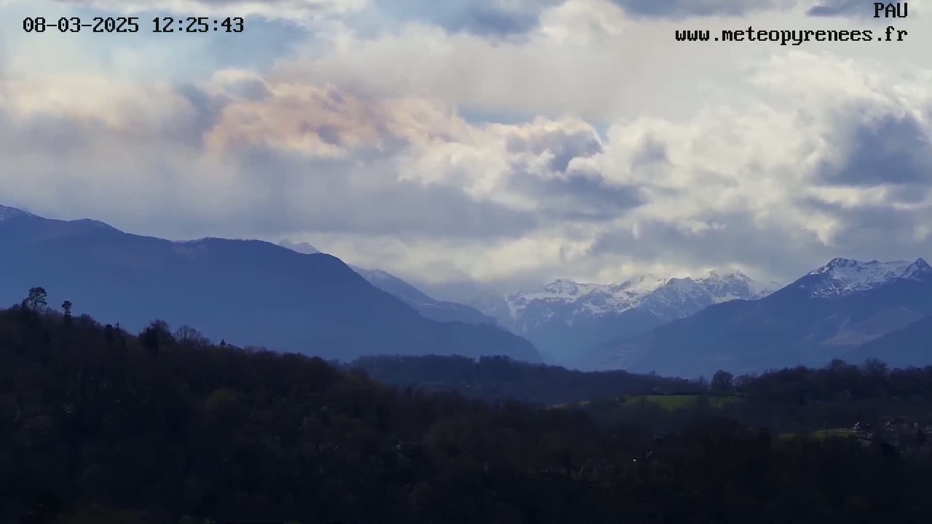 A partly cloudy sky overlooks a valley of dark green trees and hills, leading to snow-capped mountains in the distance.