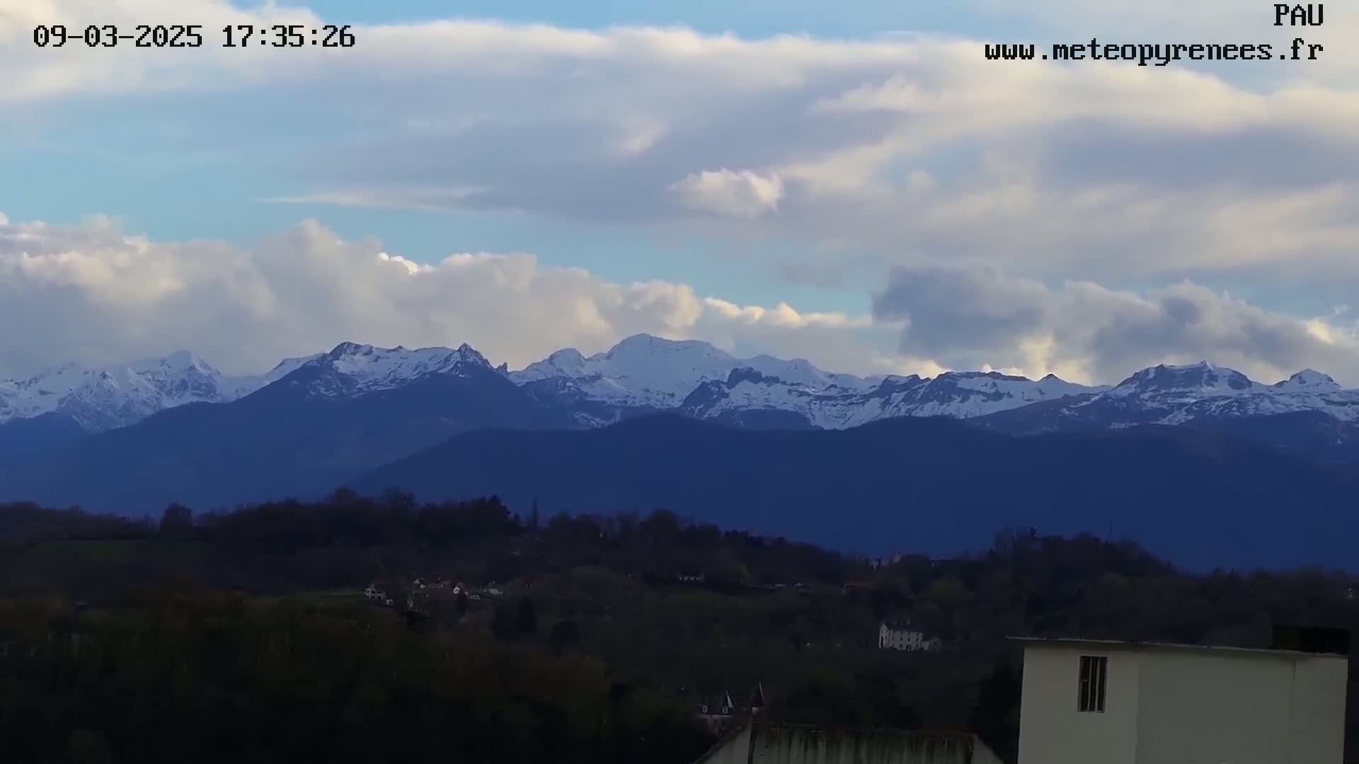 A snow-capped mountain range is visible in the distance under a partly cloudy sky, with a hilly landscape and buildings in the foreground.