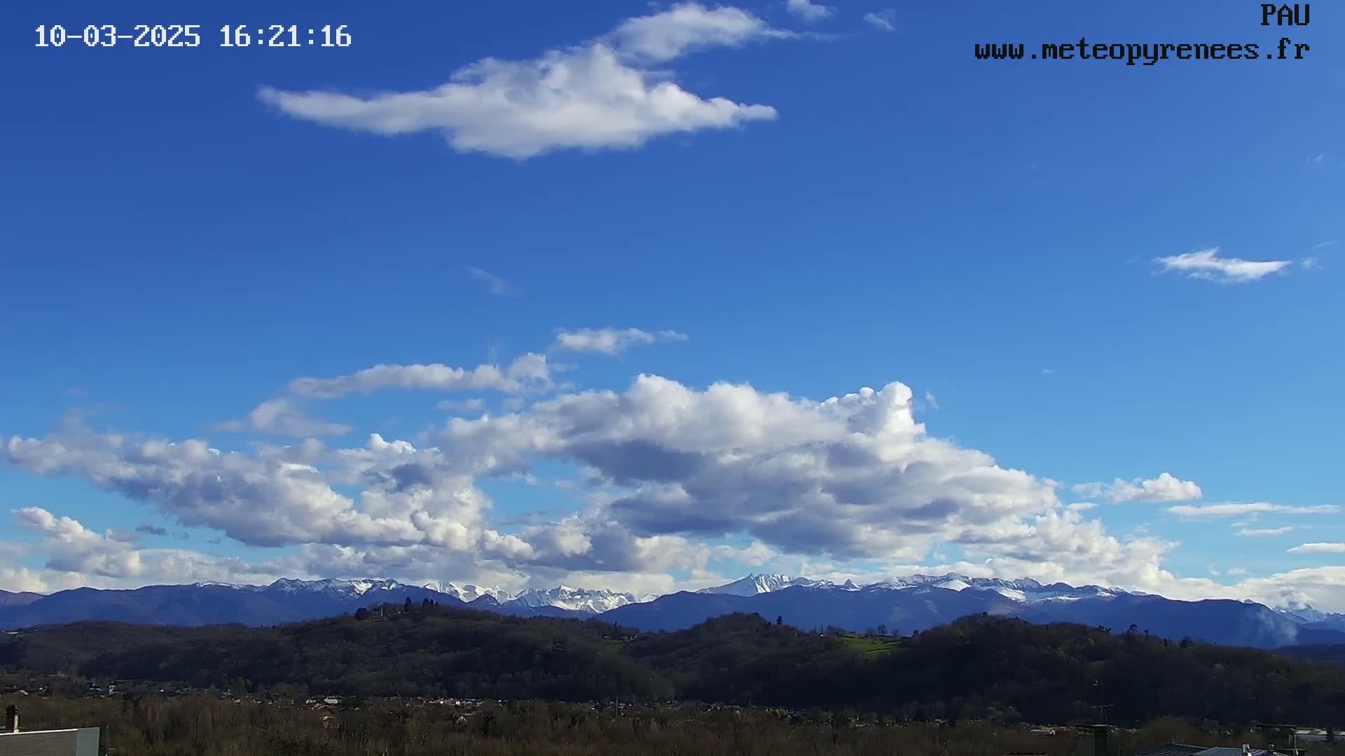 A partly cloudy blue sky overlooks a landscape of rolling hills and snow-capped mountains in the distance.