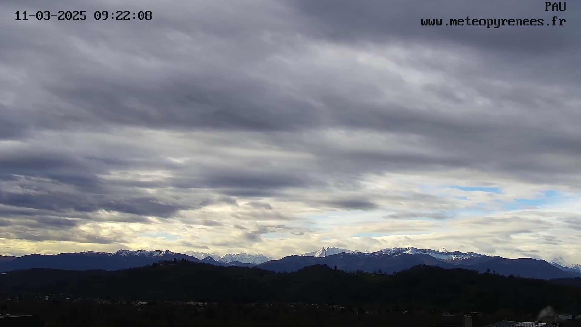A mostly cloudy sky overlooks a landscape of dark hills and distant snow-capped mountains.