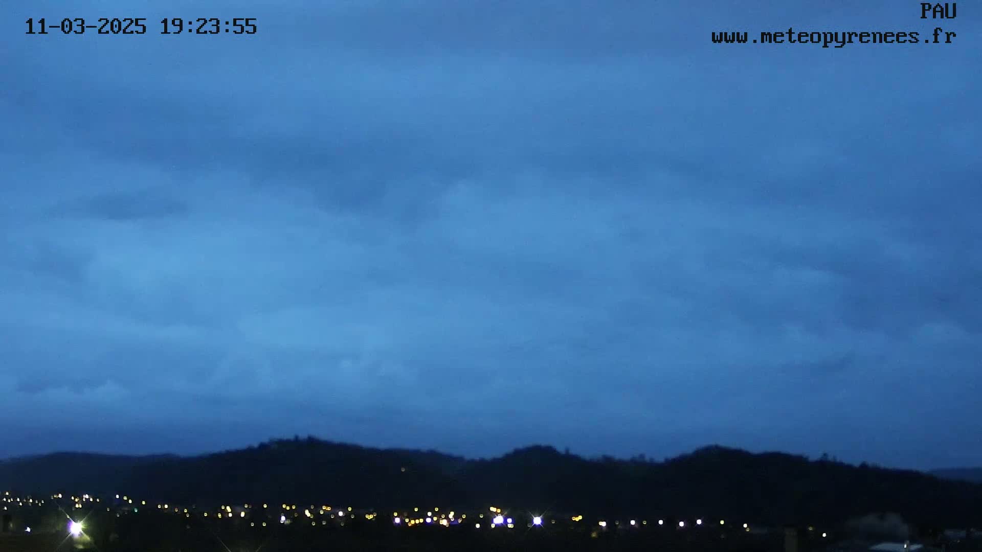 A mostly cloudy, twilight sky is over a dark, hilly landscape with a few distant lights.