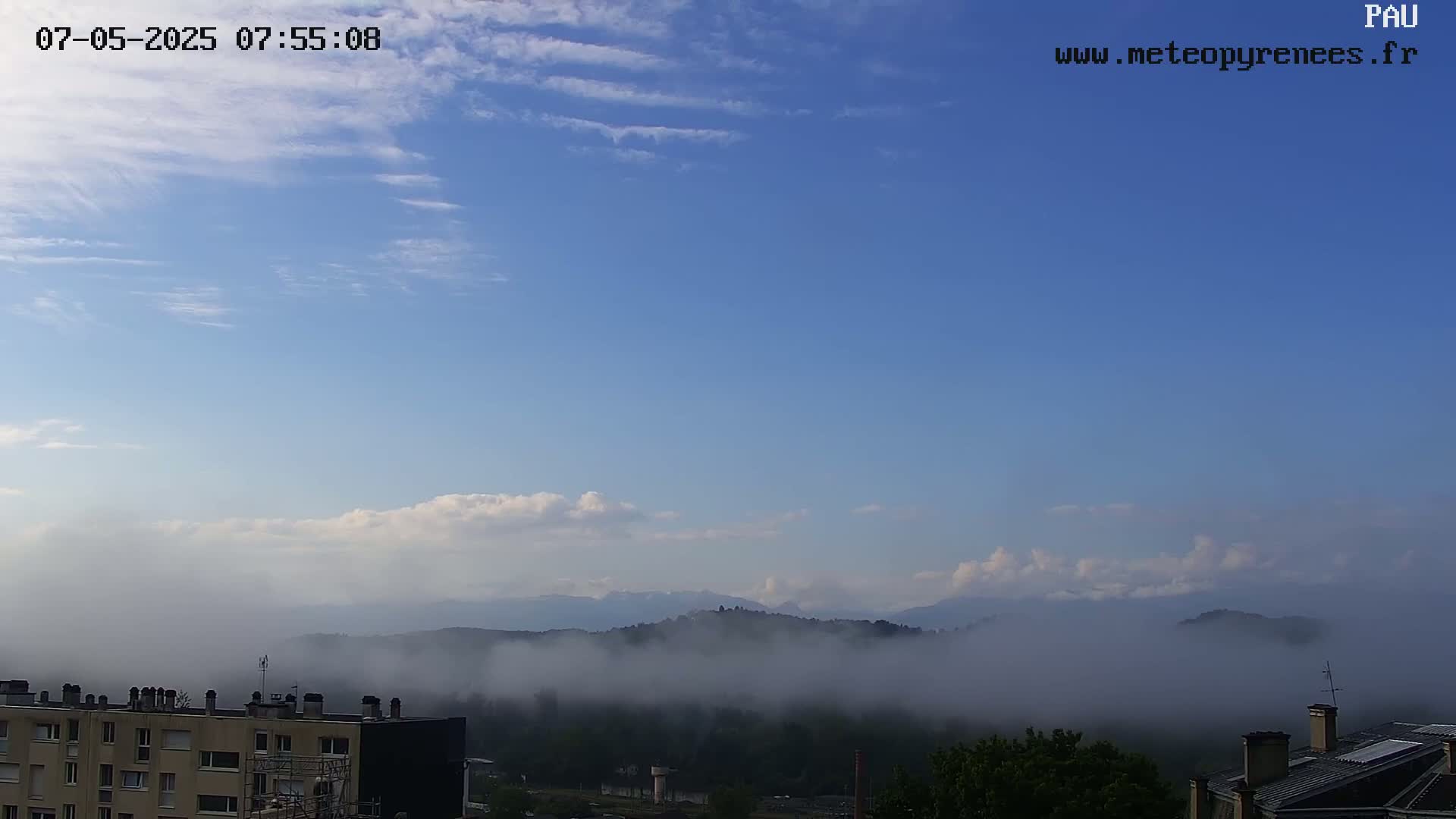 A partly cloudy sky overlooks a valley shrouded in fog, with distant mountains visible in the background and buildings in the foreground.