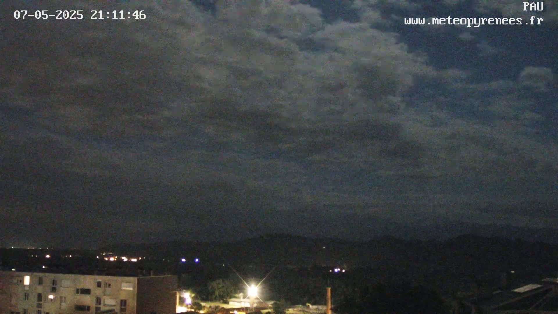 A nighttime view of a city skyline and distant hills under a mostly cloudy sky.