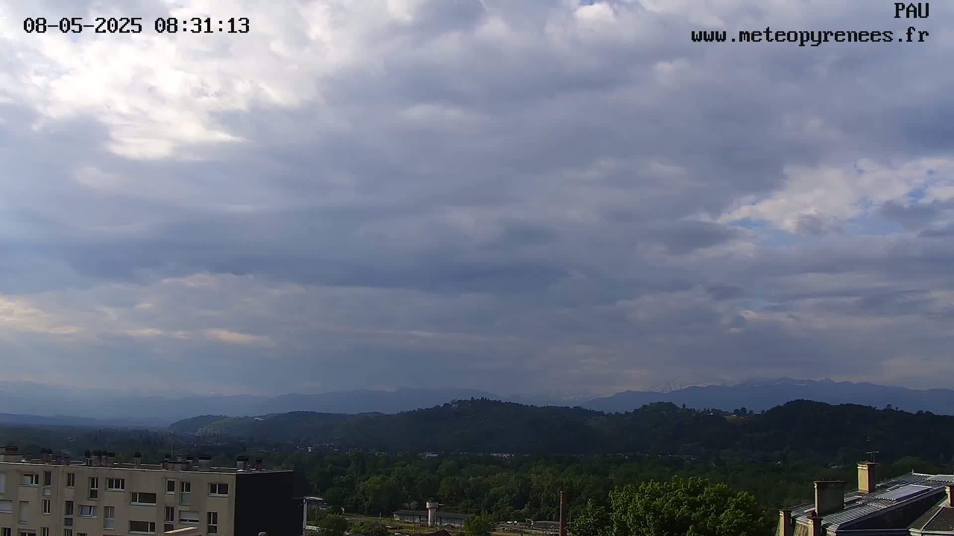 A mostly cloudy sky overlooks a valley of green trees and buildings, with mountains visible in the distance.