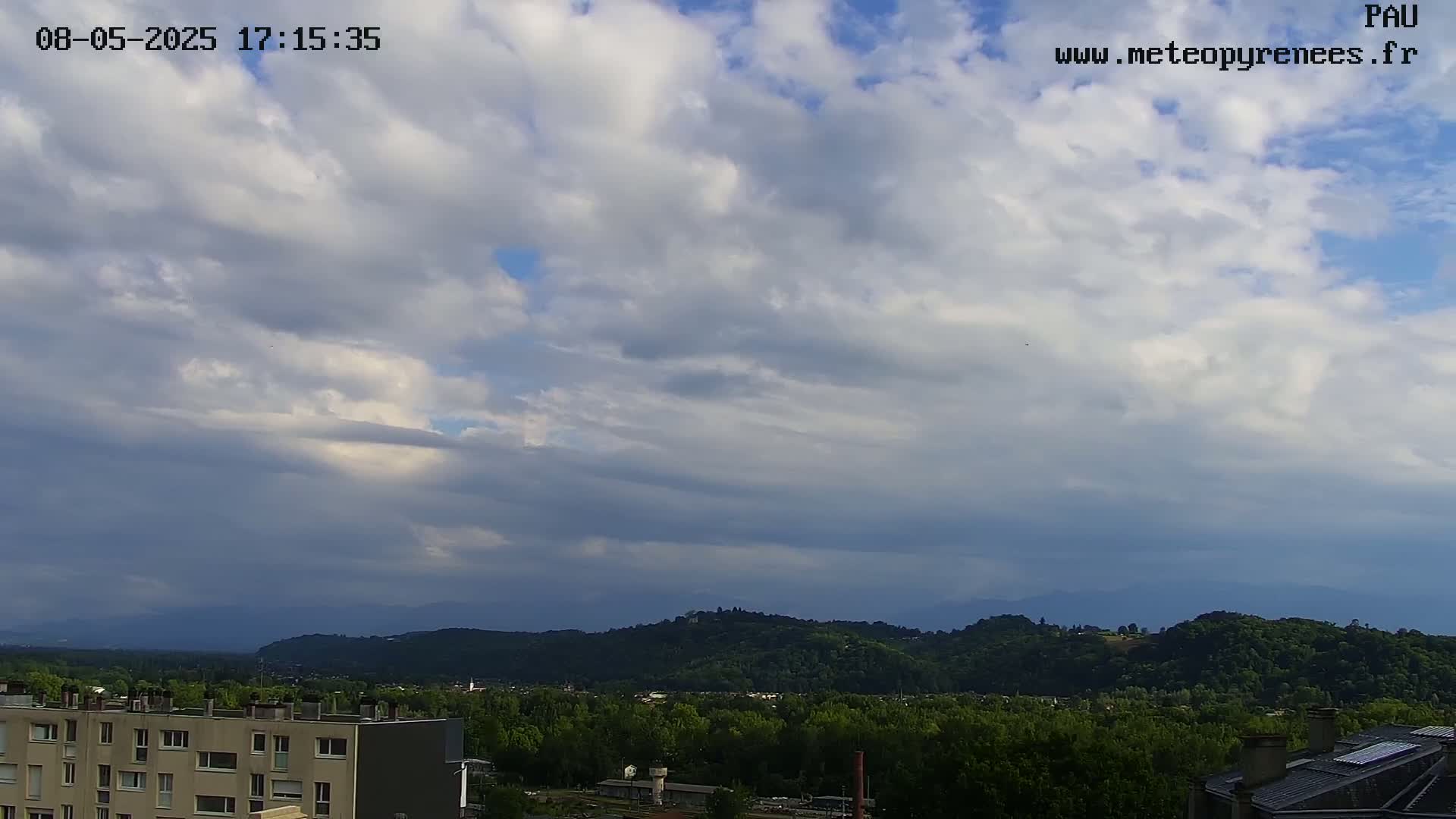 A partly cloudy sky overlooks a hilly landscape with green trees and buildings, under a mostly cloudy sky.