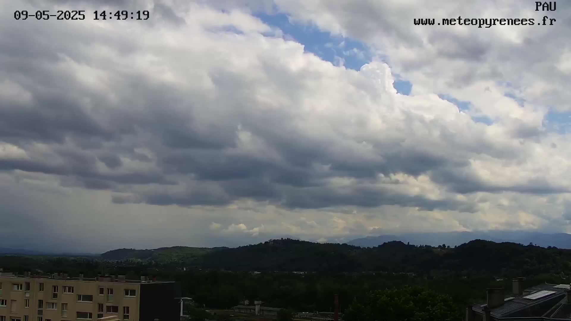 A mostly cloudy sky hangs over a hilly landscape dotted with buildings and lush green vegetation.