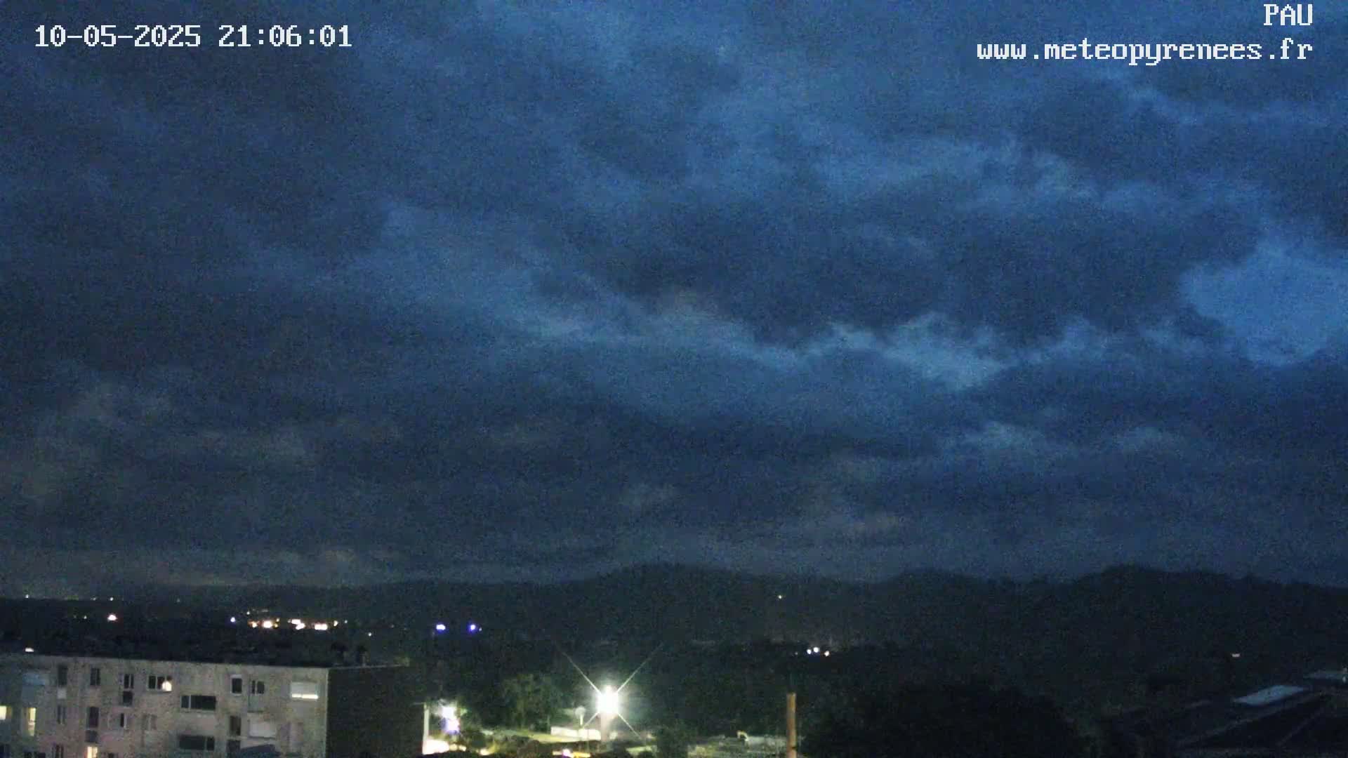 A nighttime view of a city with dark, cloudy skies and hills in the distance.