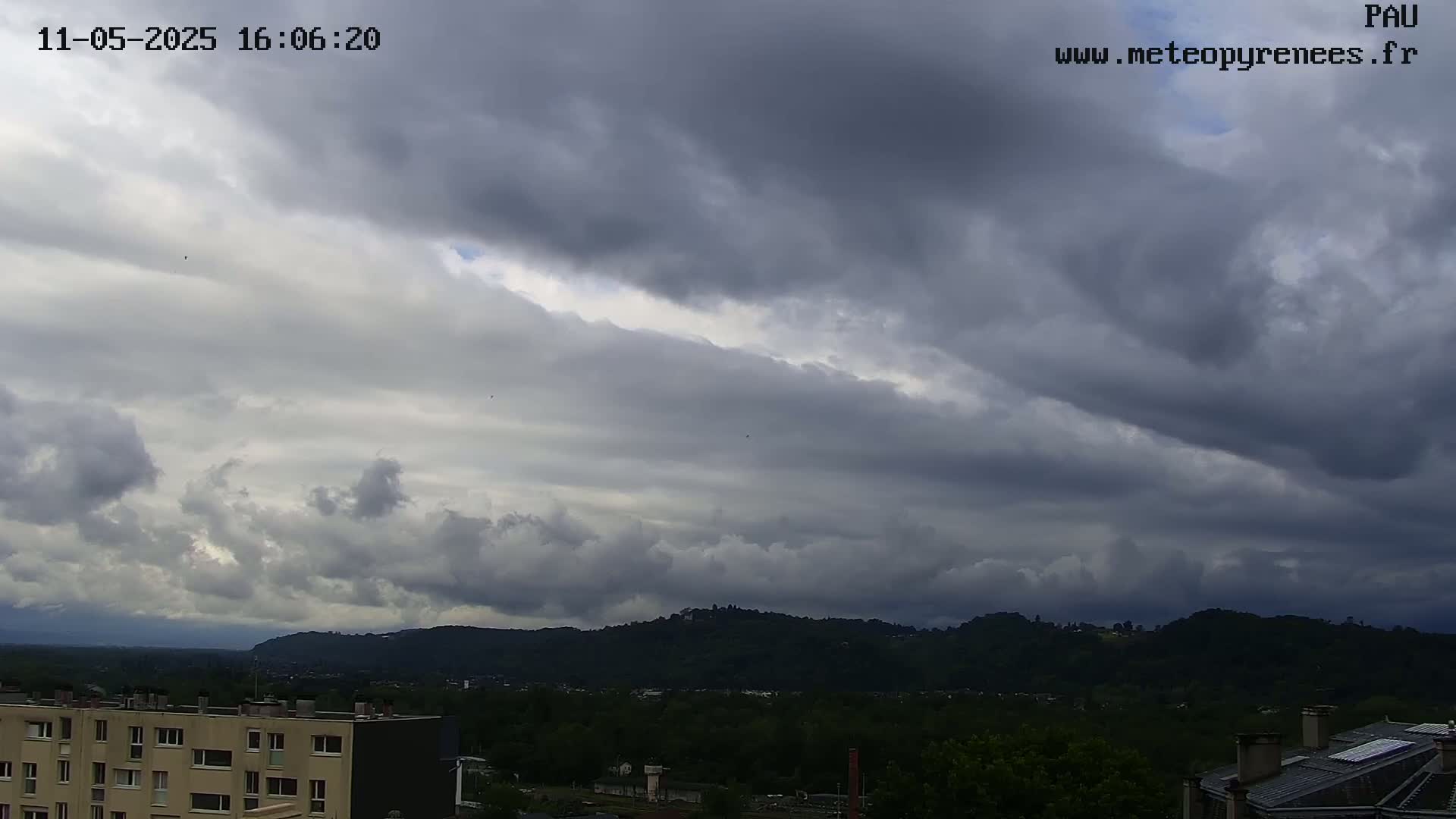 Overcast skies with dark, heavy clouds loom over a city nestled in a valley surrounded by dark green hills.