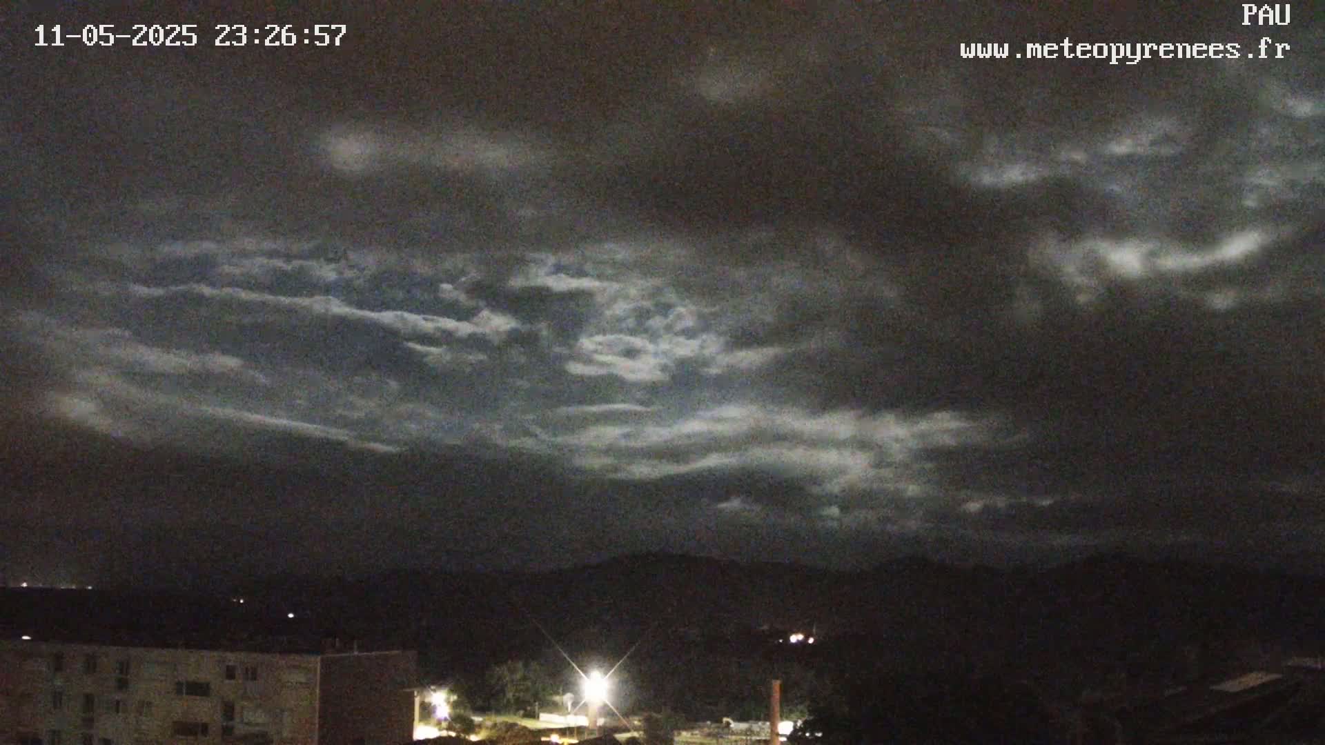 A nighttime view of a partly cloudy sky over dark mountains and a town with buildings and lights.