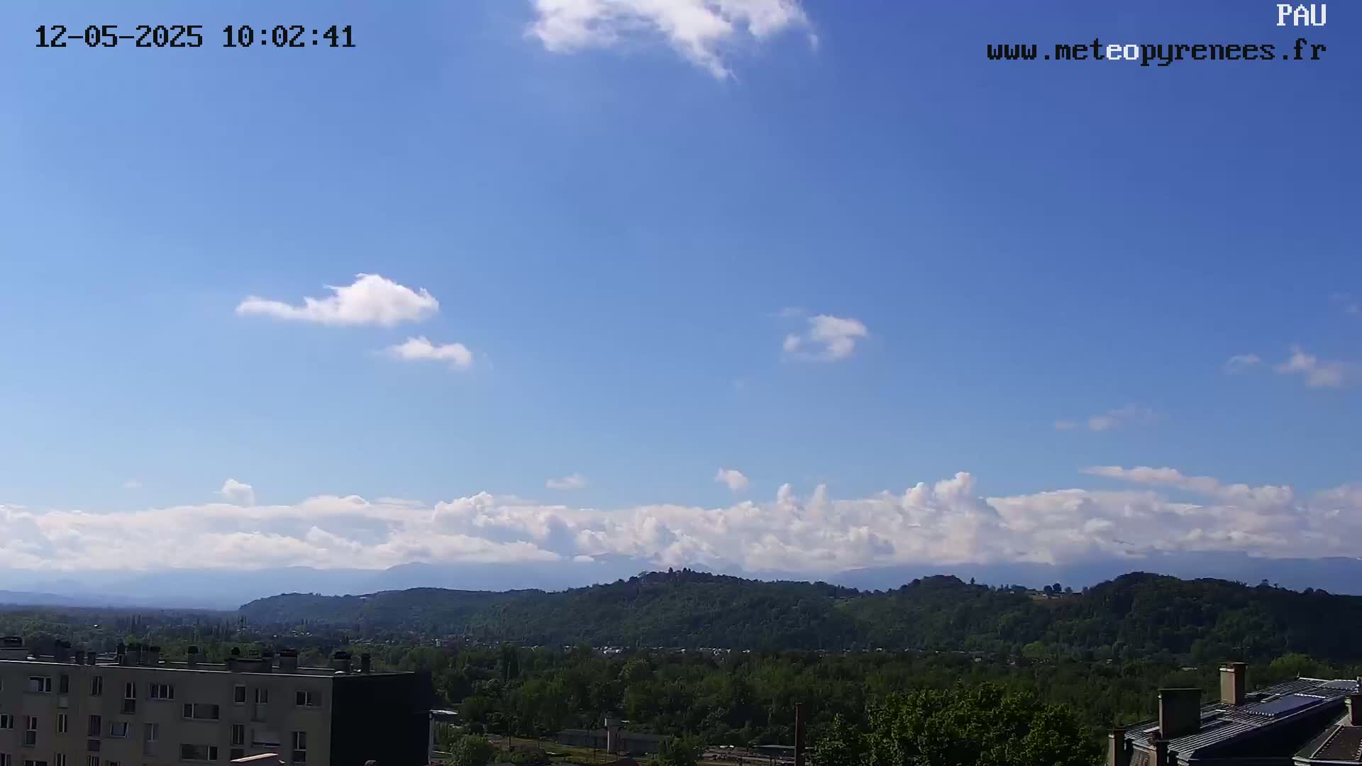 A partly cloudy sky overlooks a hilly landscape with green trees and buildings, under a mostly clear blue sky.