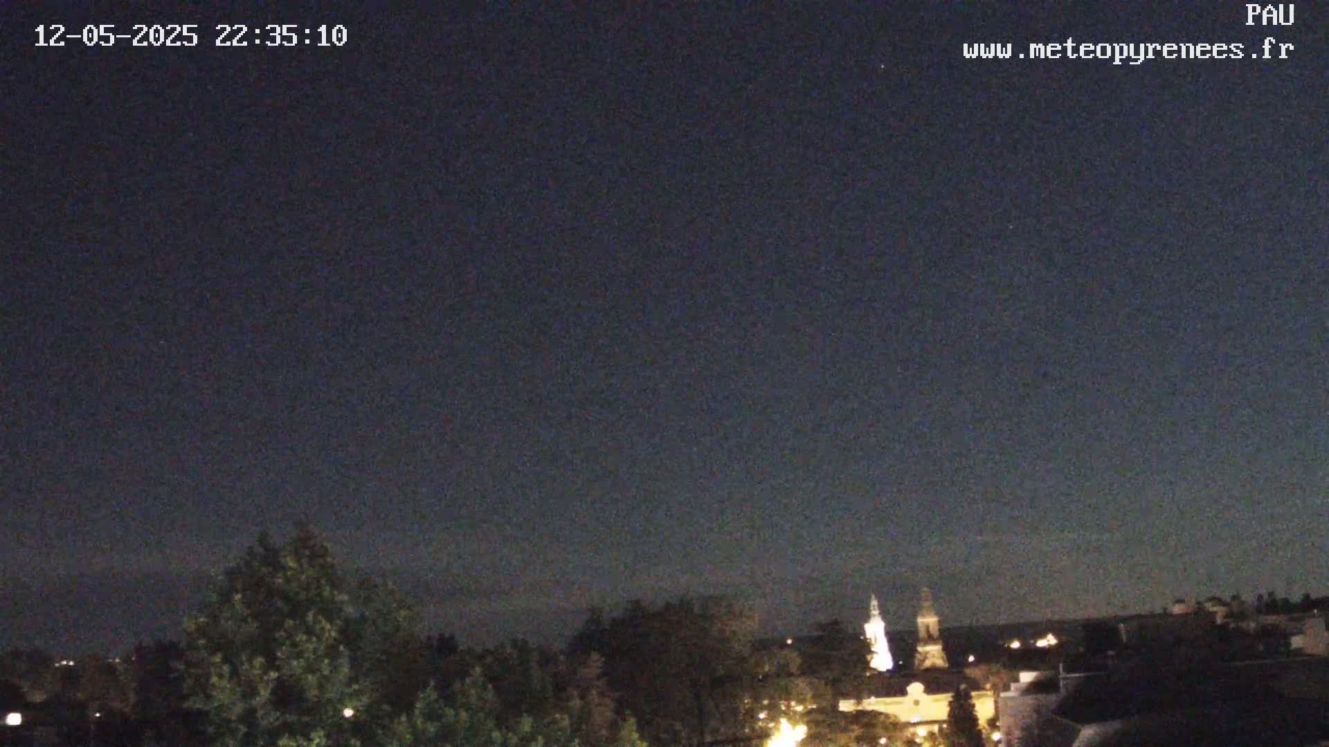 A nighttime view of a town with a church steeple, partially obscured by trees under a clear, dark sky.