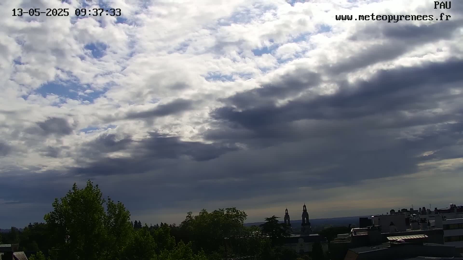 A partly cloudy sky hangs over a cityscape with green trees and a pair of church steeples visible in the distance.