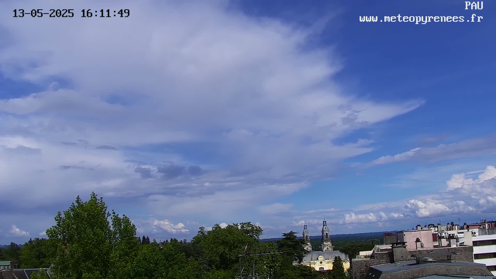 A partly cloudy sky with puffy white clouds against a blue background overlooks a town with green trees and buildings.