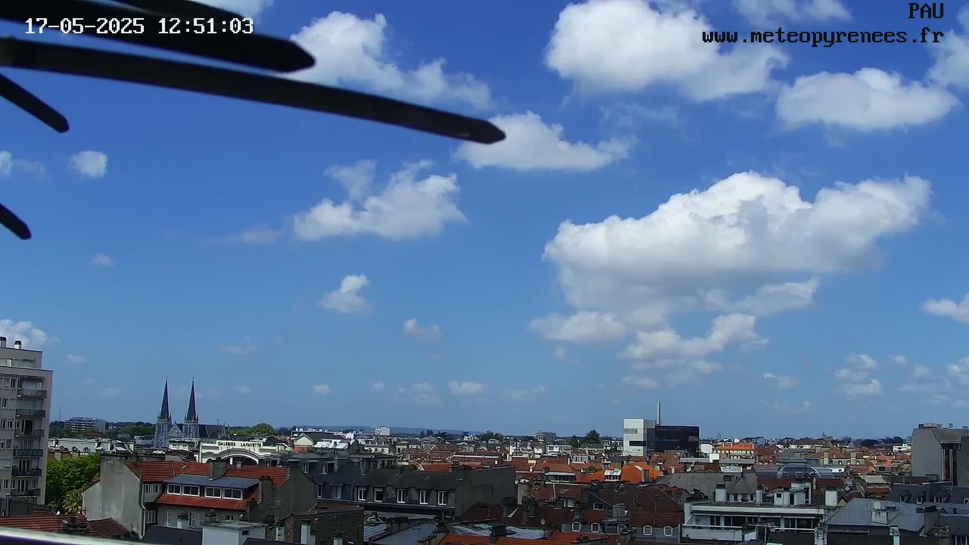 A mostly sunny sky with scattered cumulus clouds overlooks a cityscape of red-tiled roofs and taller buildings.