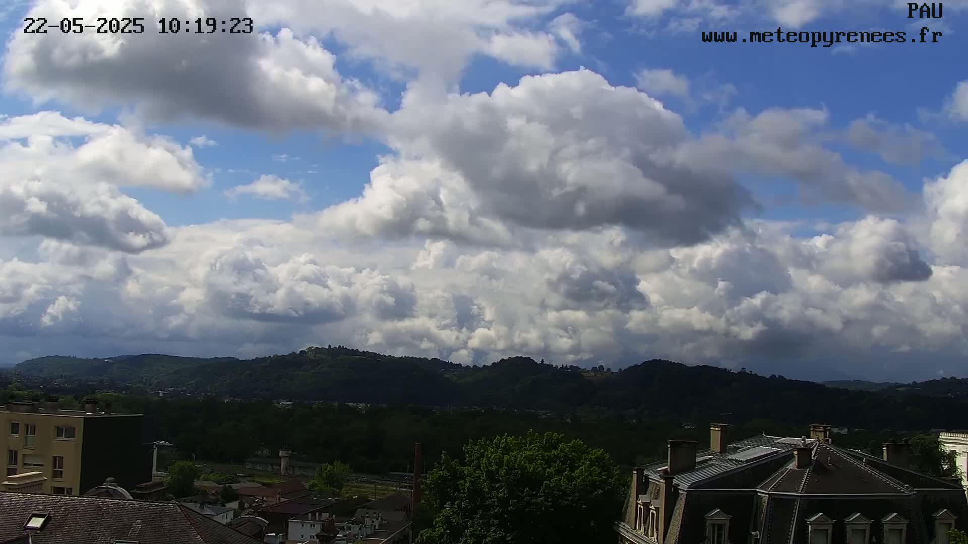 A partly cloudy sky over a hilly landscape with green trees and buildings.