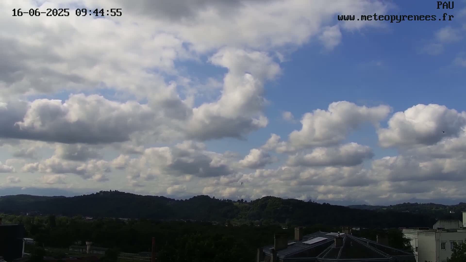 A partly cloudy sky over a landscape of rolling green hills and a town with buildings, seen from an elevated viewpoint.