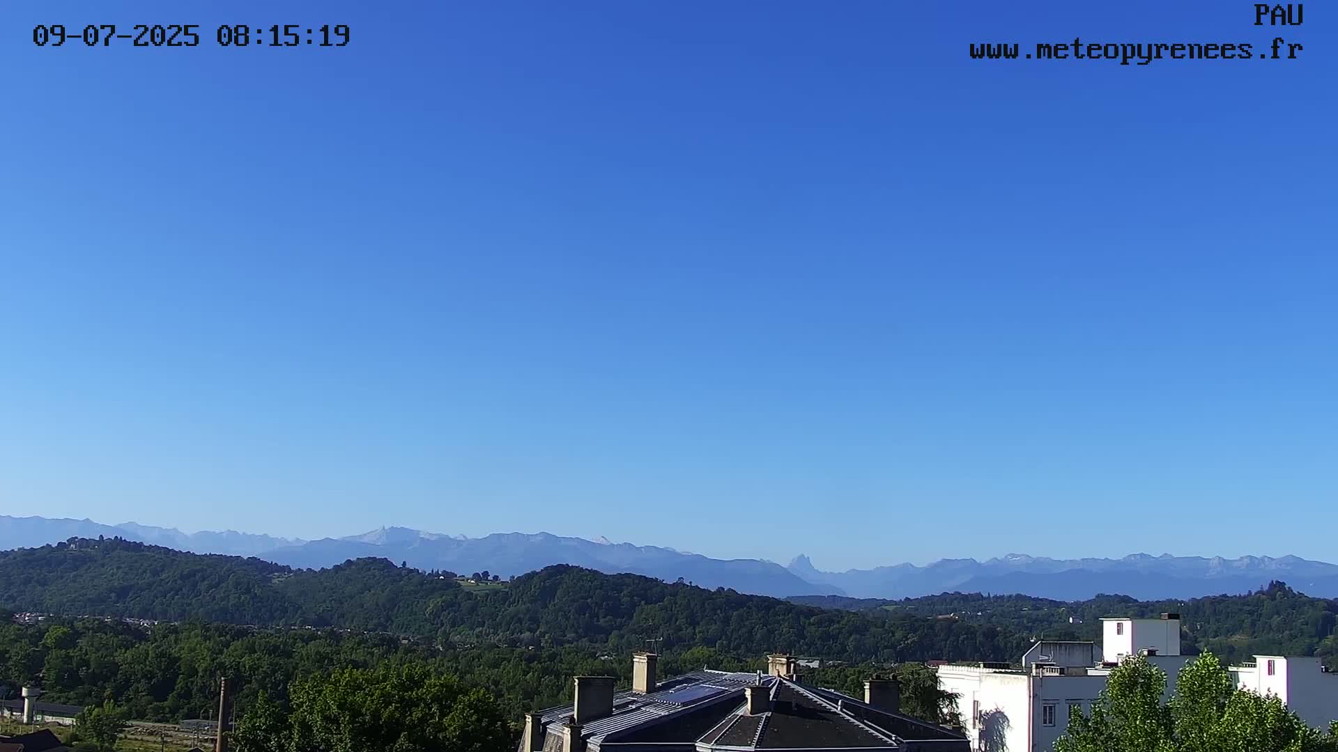 A clear blue sky overlooks a landscape of green hills and distant mountains, with rooftops of buildings visible in the foreground.