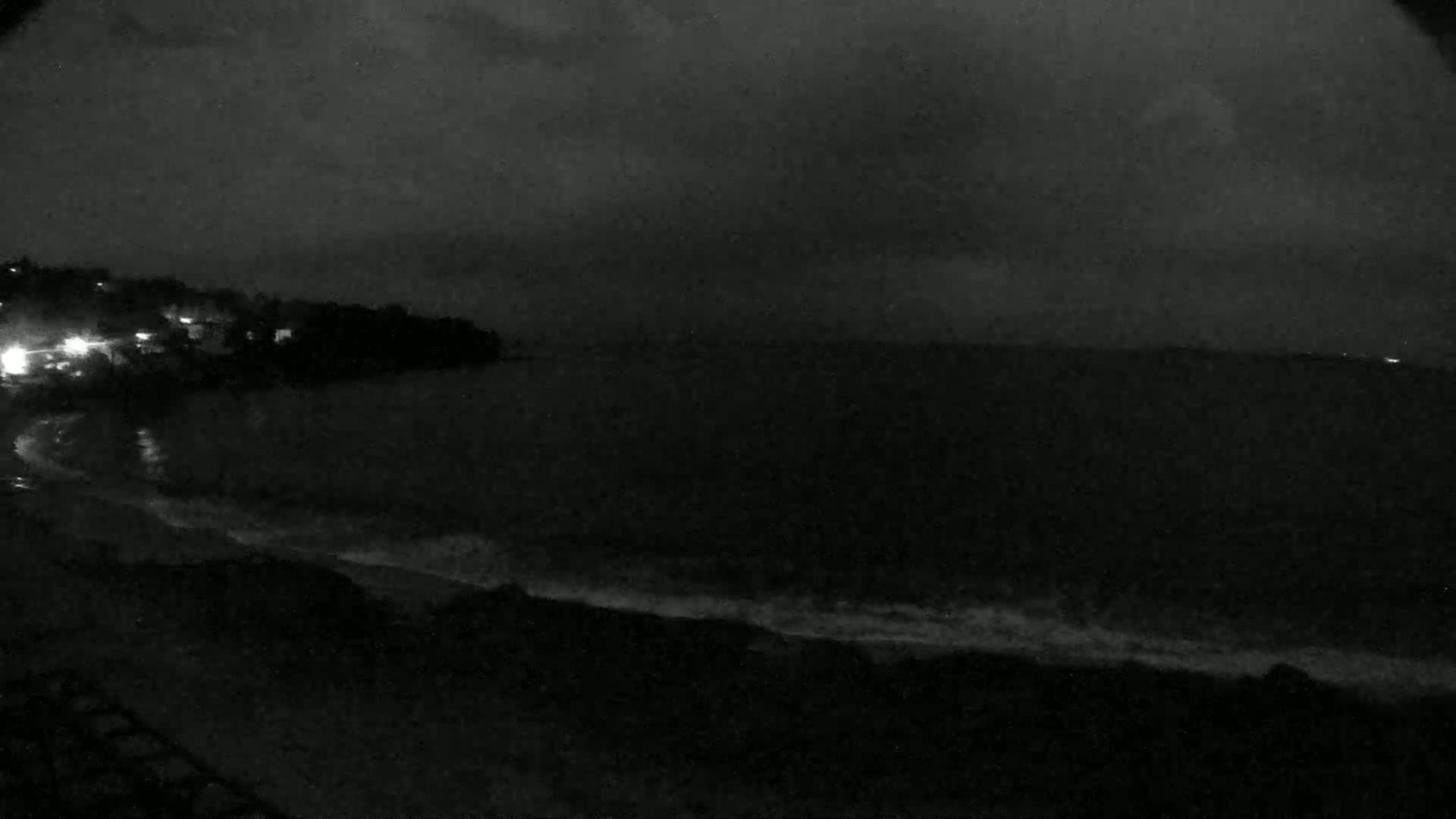 A nighttime, dark, overcast coastal scene shows a shoreline with a few lights visible in the distance, and gentle waves lapping the shore.