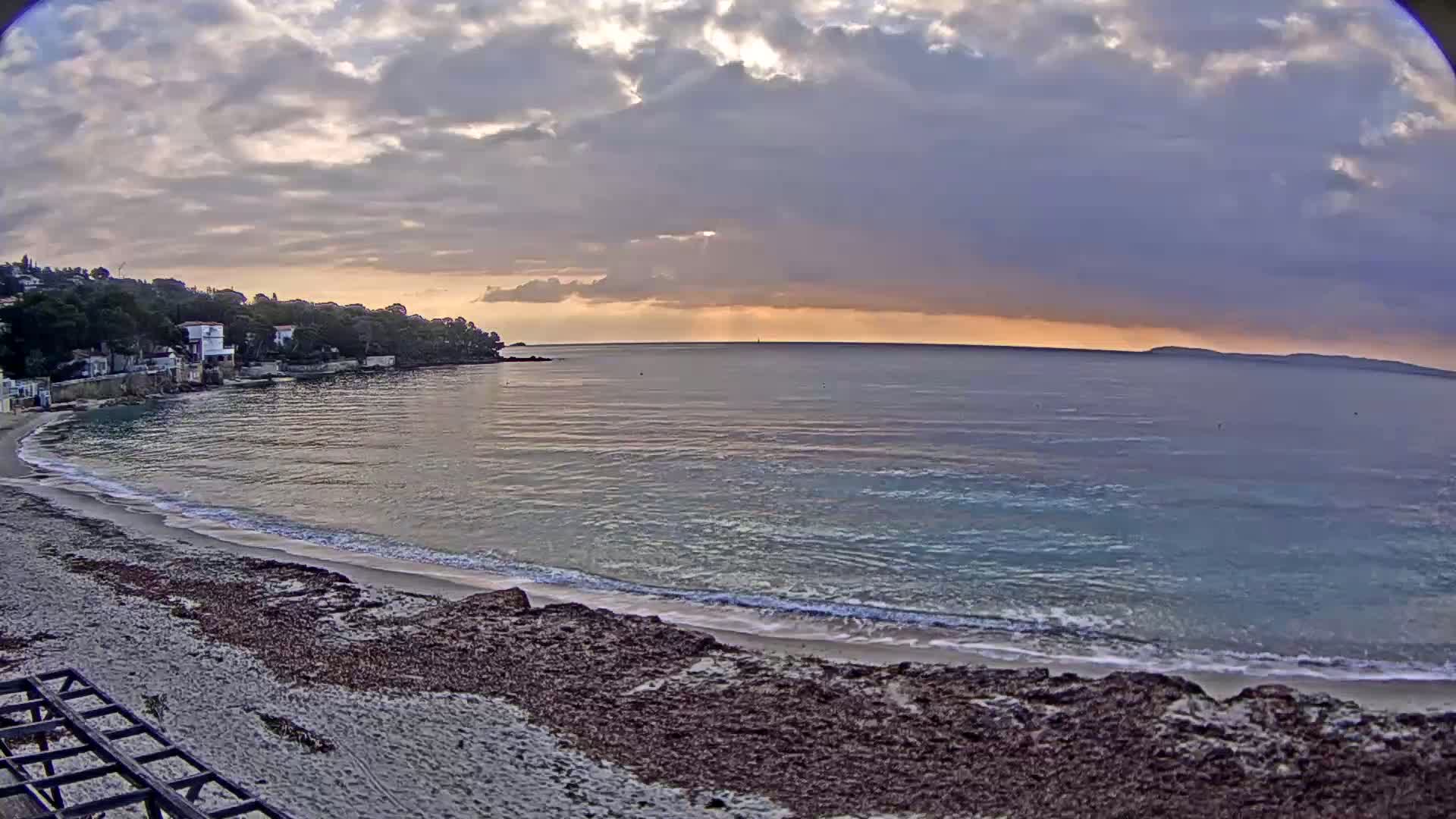 A sandy beach with dark seaweed lines the edge of a calm sea under a cloudy, sunrise sky.