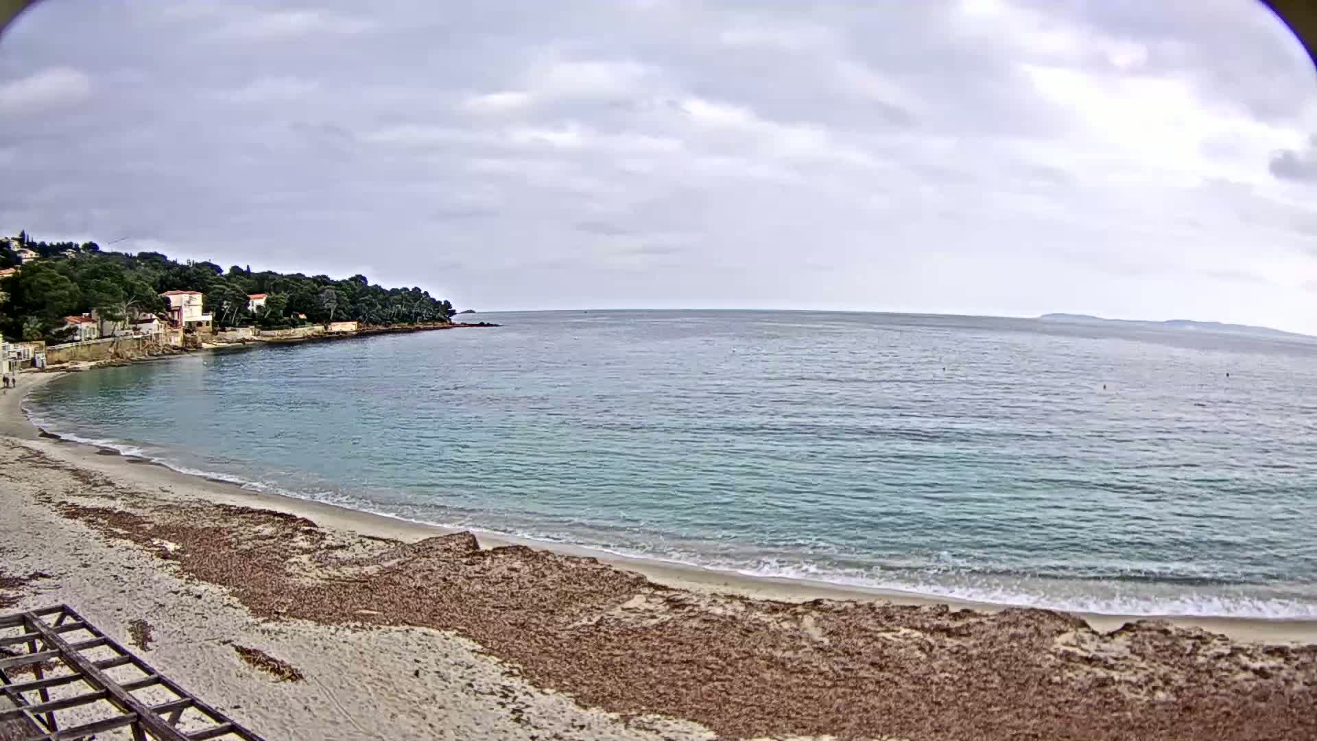 A sandy beach with seaweed lines the calm, turquoise water of a bay under a cloudy sky, with houses and trees visible on the far shore.