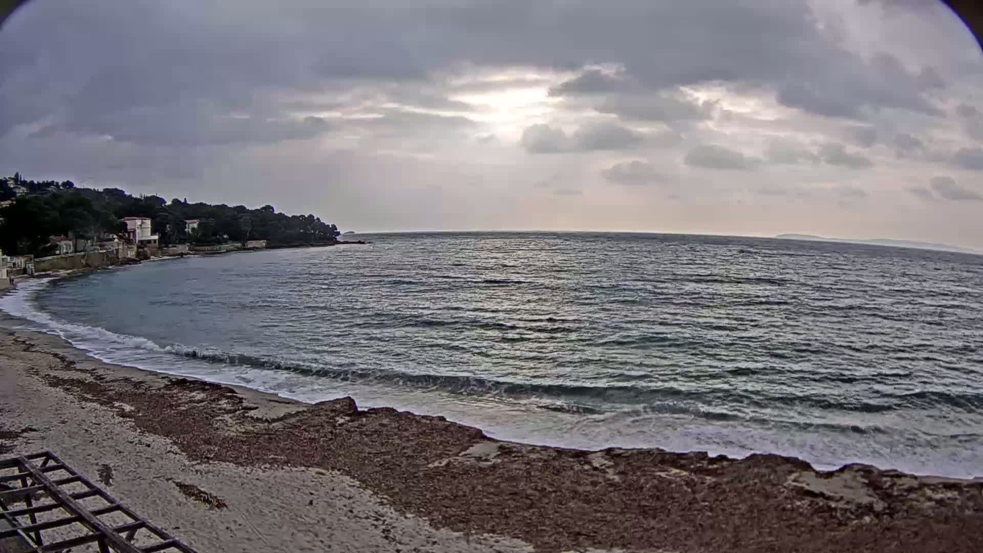 A sandy beach with small waves laps against a rocky shore, under a mostly cloudy sky with a hint of sunlight.