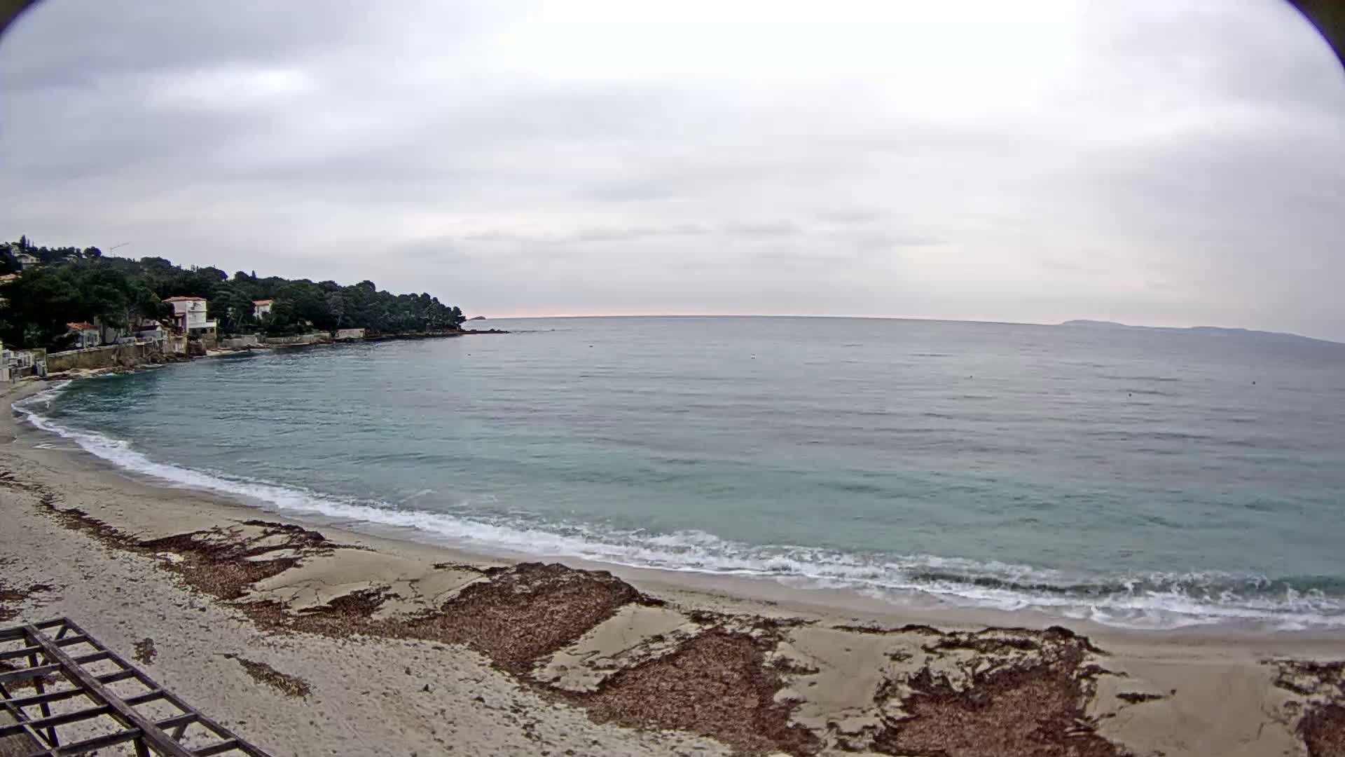 A sandy beach with small waves laps the shore under an overcast sky, with houses and trees visible on a hillside in the background.