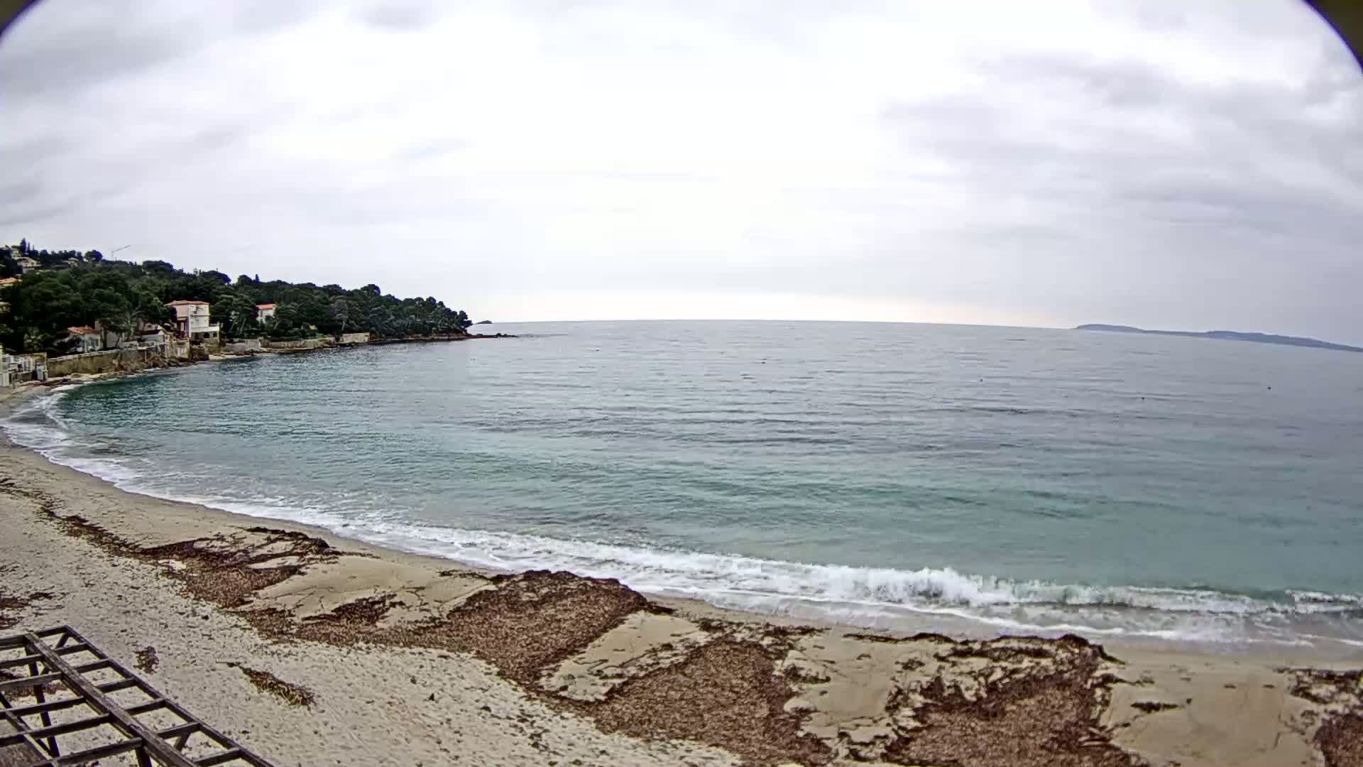 A sandy beach with seaweed washes gently onto the shore of a calm, teal ocean under an overcast sky, with houses and trees visible on a hillside in the background.