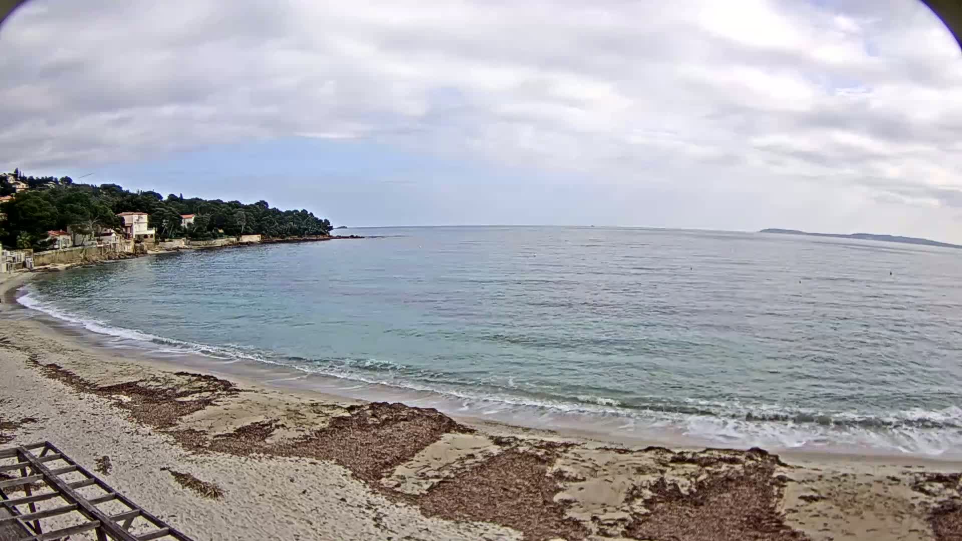 A sandy beach with seaweed washes up on the shore next to calm, light-blue water under an overcast sky.