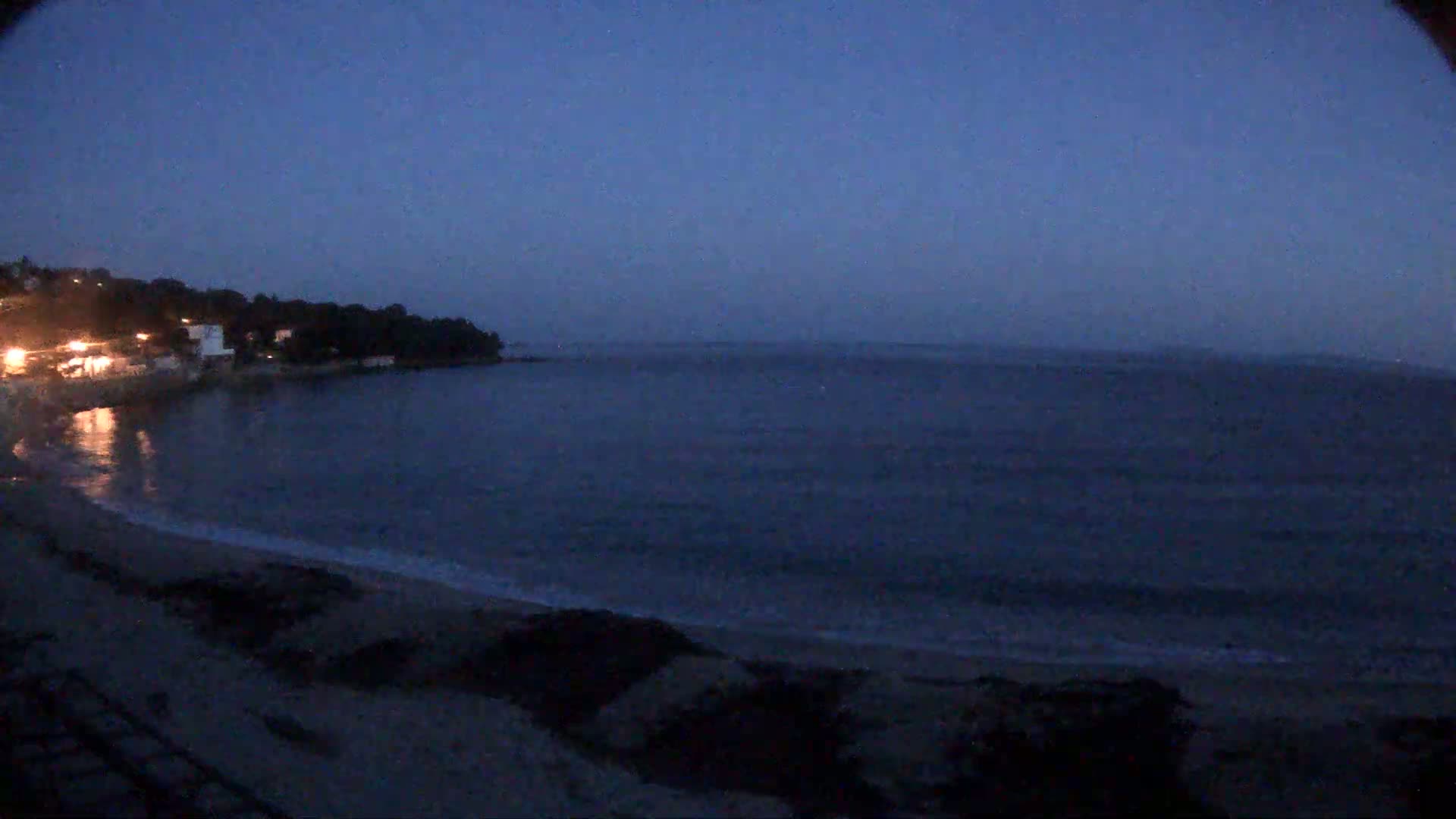 A dark, mostly clear night shows a calm, dark-blue ocean lapping a sandy beach, with some lights visible on the distant shore.