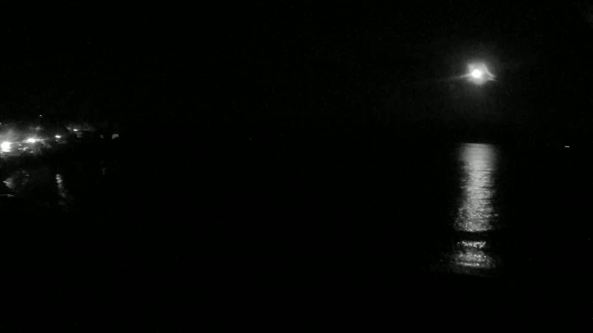 A bright moon illuminates a dark body of water at night, with a faint light source visible on the left.