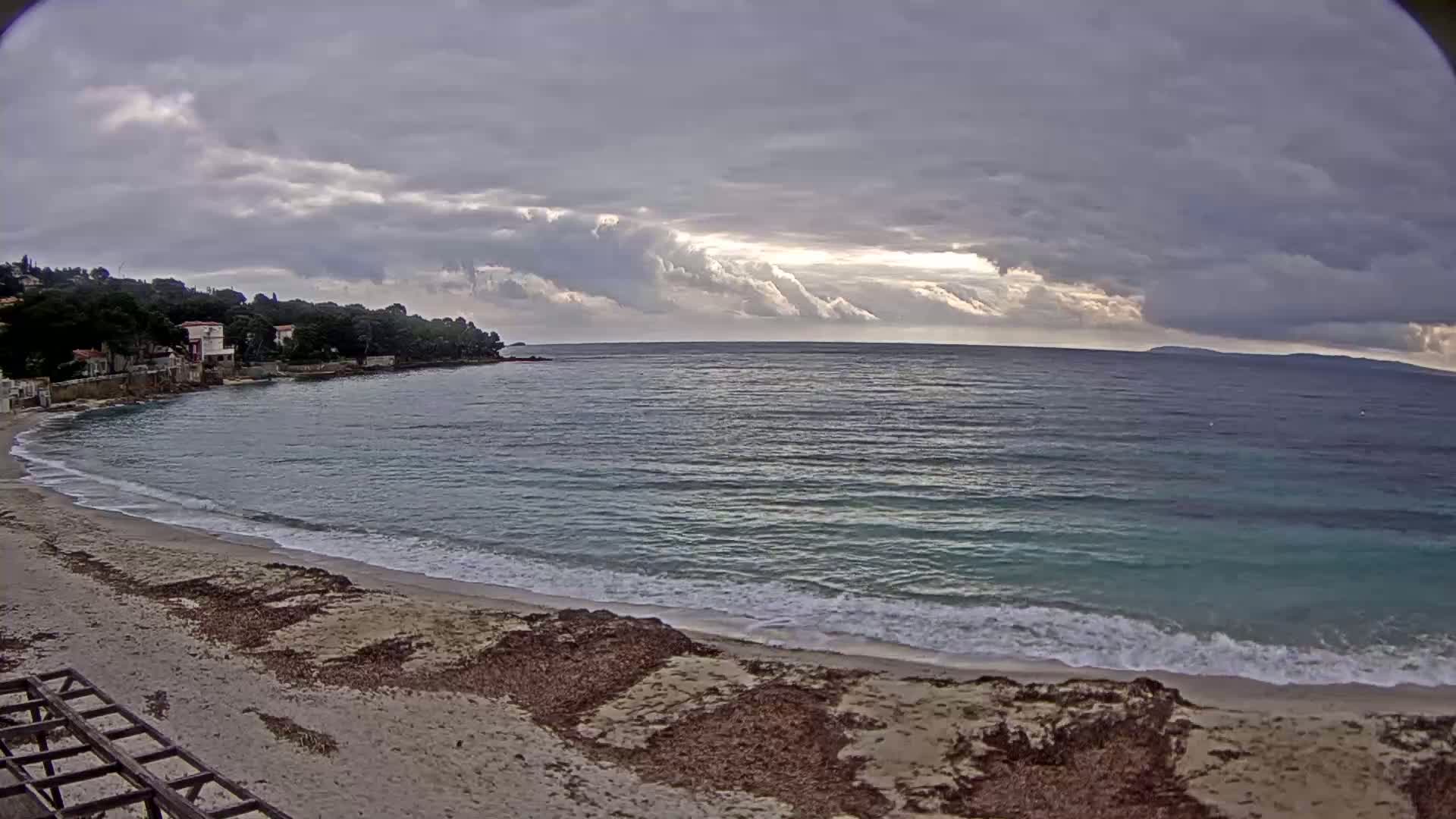 A sandy beach with small waves laps the shore under a cloudy sky, with a calm sea extending to a distant shoreline.