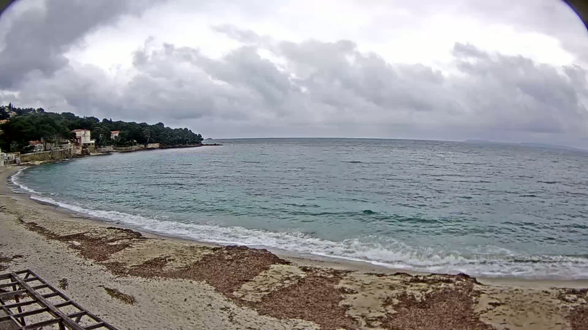 A sandy beach with small waves laps the shore under a cloudy sky, with houses and trees visible on a nearby hill.