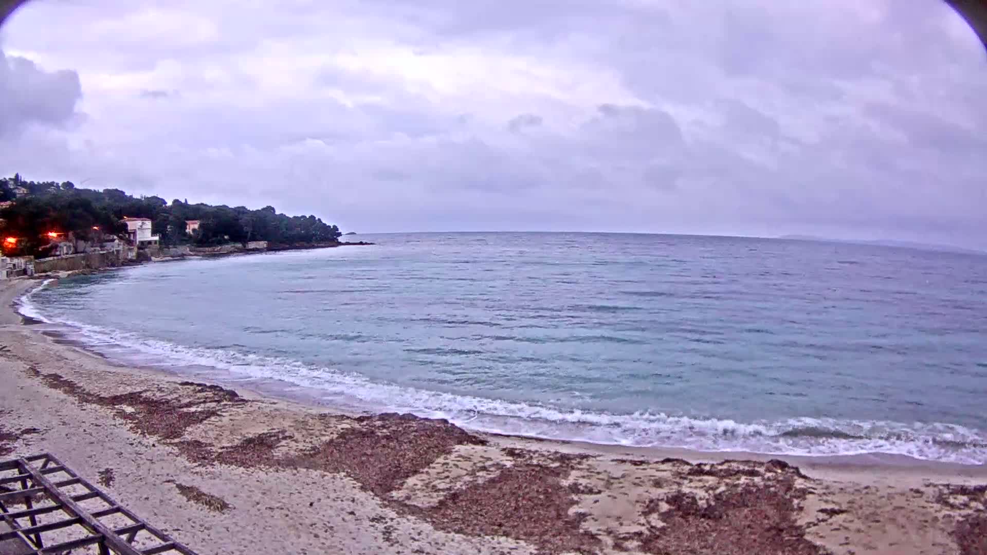 A sandy beach with seaweed lines the edge of a calm, light-blue sea under an overcast sky.
