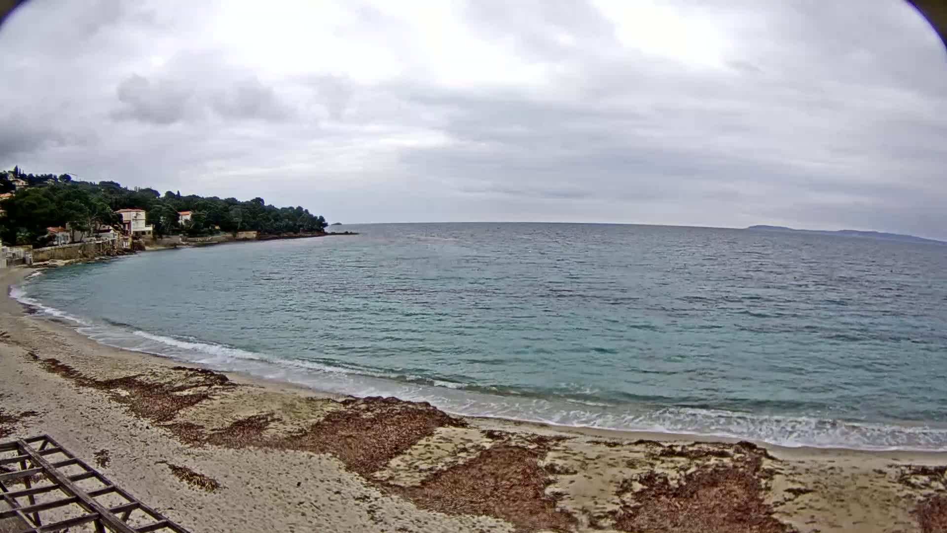 A sandy beach with seaweed and small waves laps the shore of a calm, teal ocean under a cloudy sky, with houses and trees visible on a nearby hill.