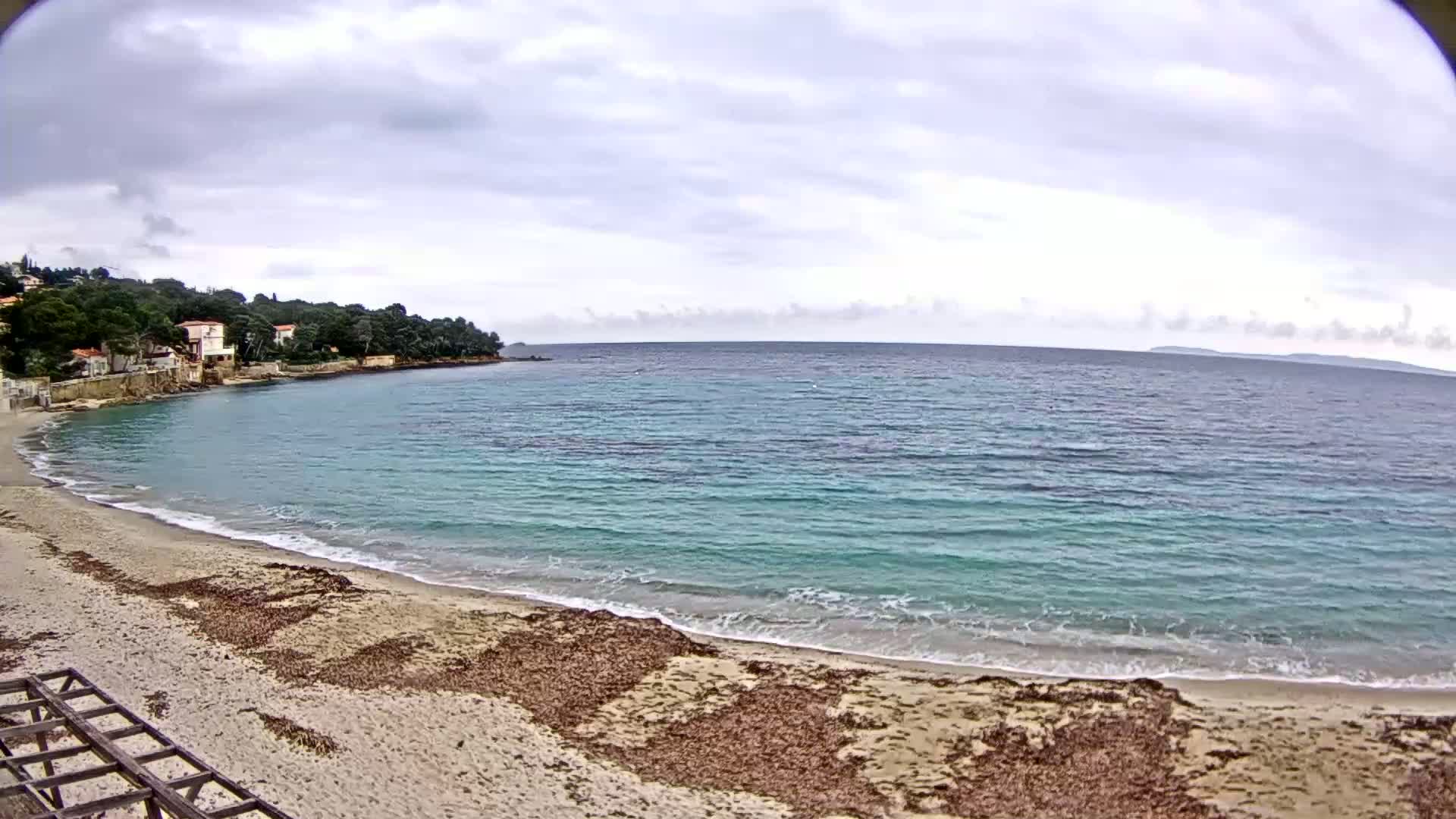 A sandy beach with calm, turquoise water laps gently at the shore under an overcast sky, backed by a line of houses nestled amongst green trees.