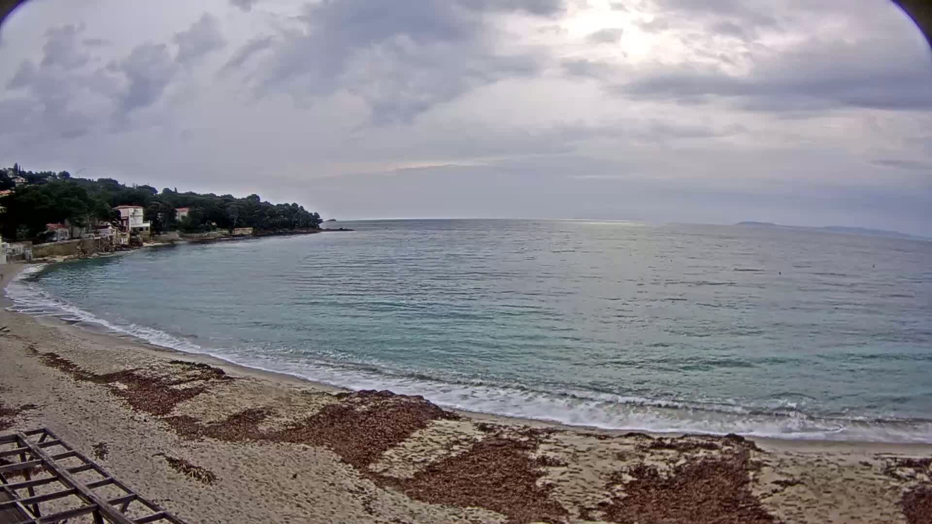 A sandy beach with small waves laps the shore under an overcast sky, with a line of houses nestled amongst trees visible in the distance.