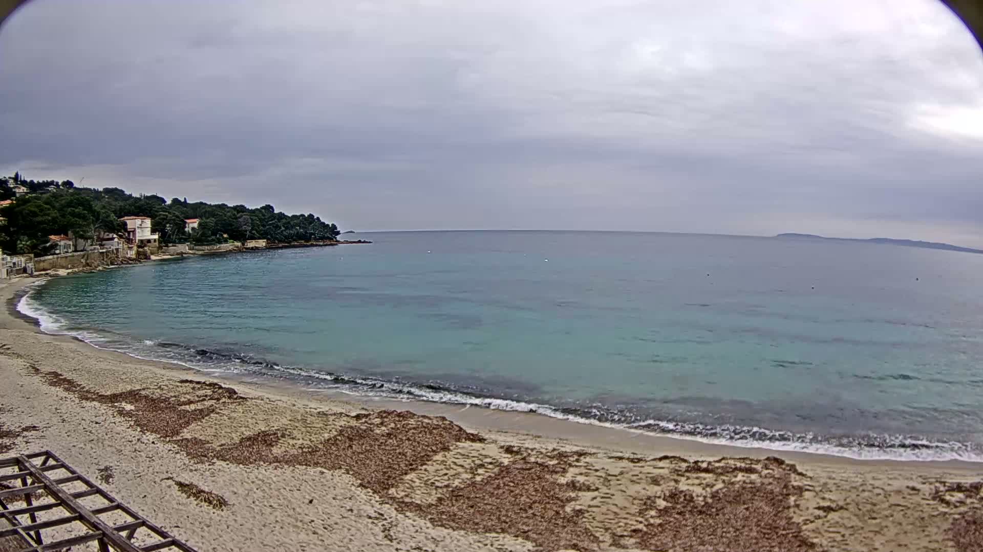 A sandy beach with calm, turquoise water under a cloudy sky, bordered by houses and trees.