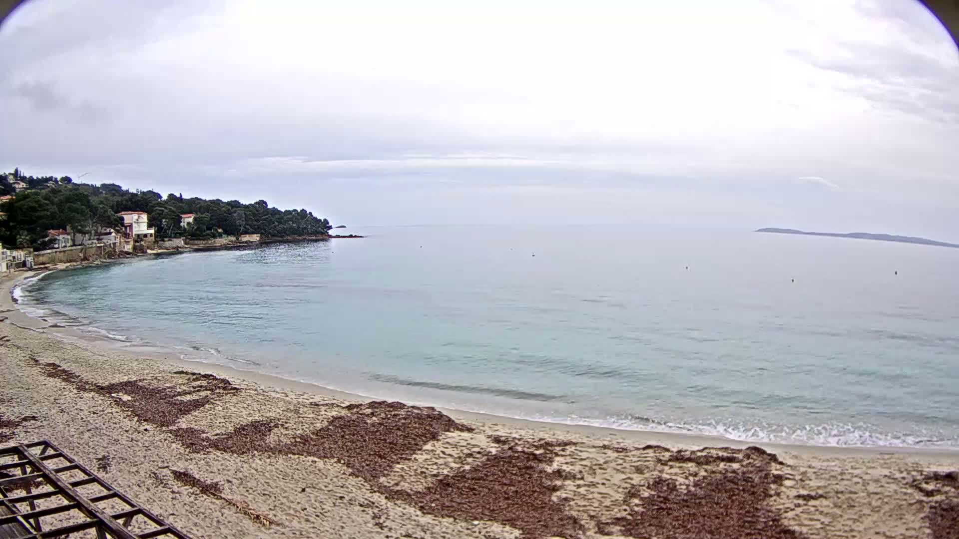 A sandy beach curves around a calm, light-blue bay under an overcast sky, with houses and trees visible on the distant headland.