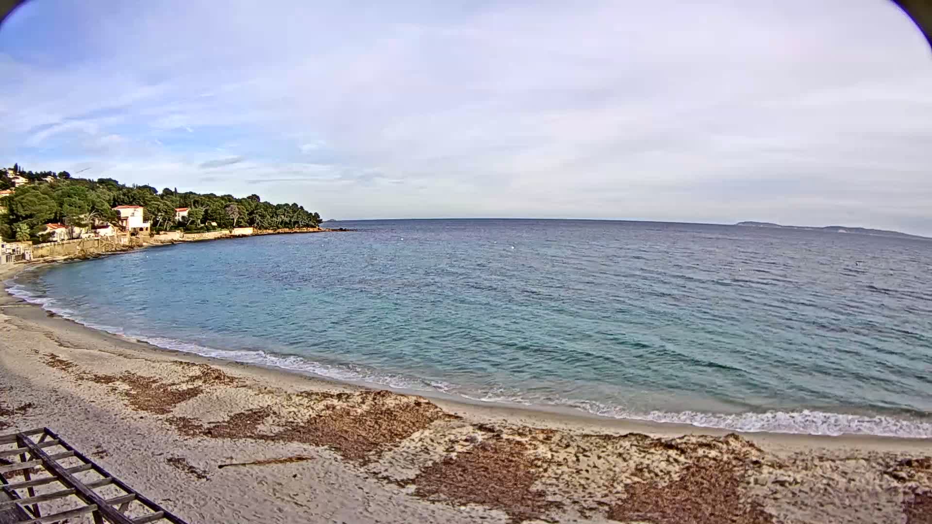 A sandy beach curves into a calm, turquoise sea under a partly cloudy sky, backed by a line of houses nestled amongst green trees on a hill.