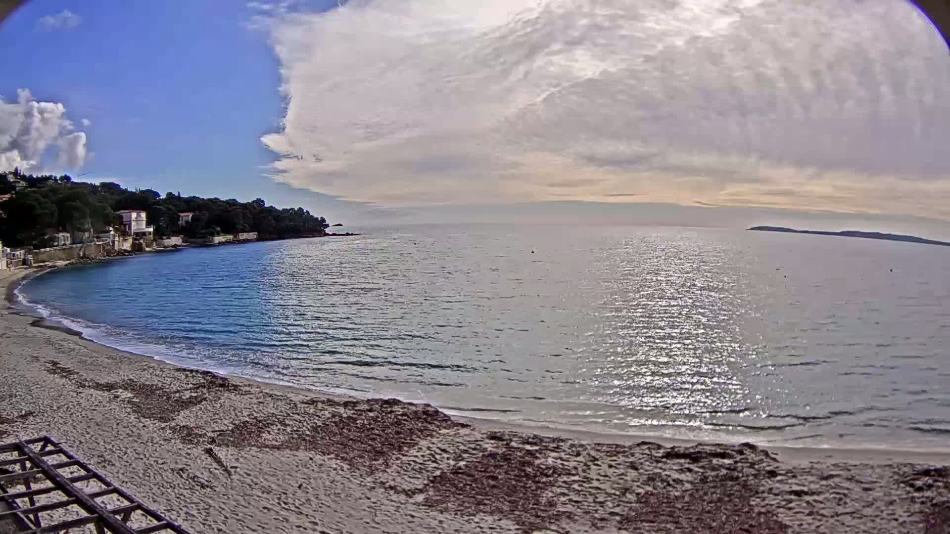 A sandy beach curves around a calm bay with blue water under a partly cloudy sky.