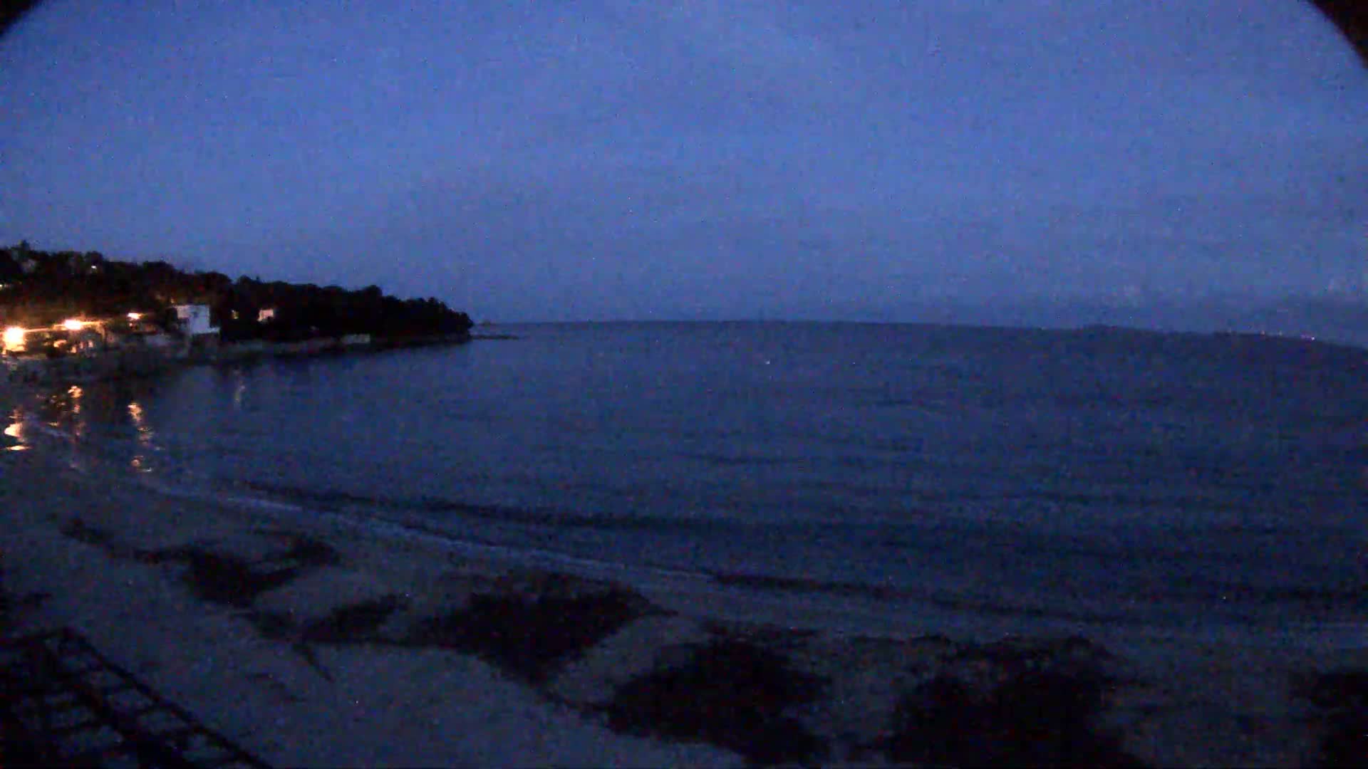A dark, calm sea laps a sandy shore at twilight, with a line of trees and buildings visible on the coast.
