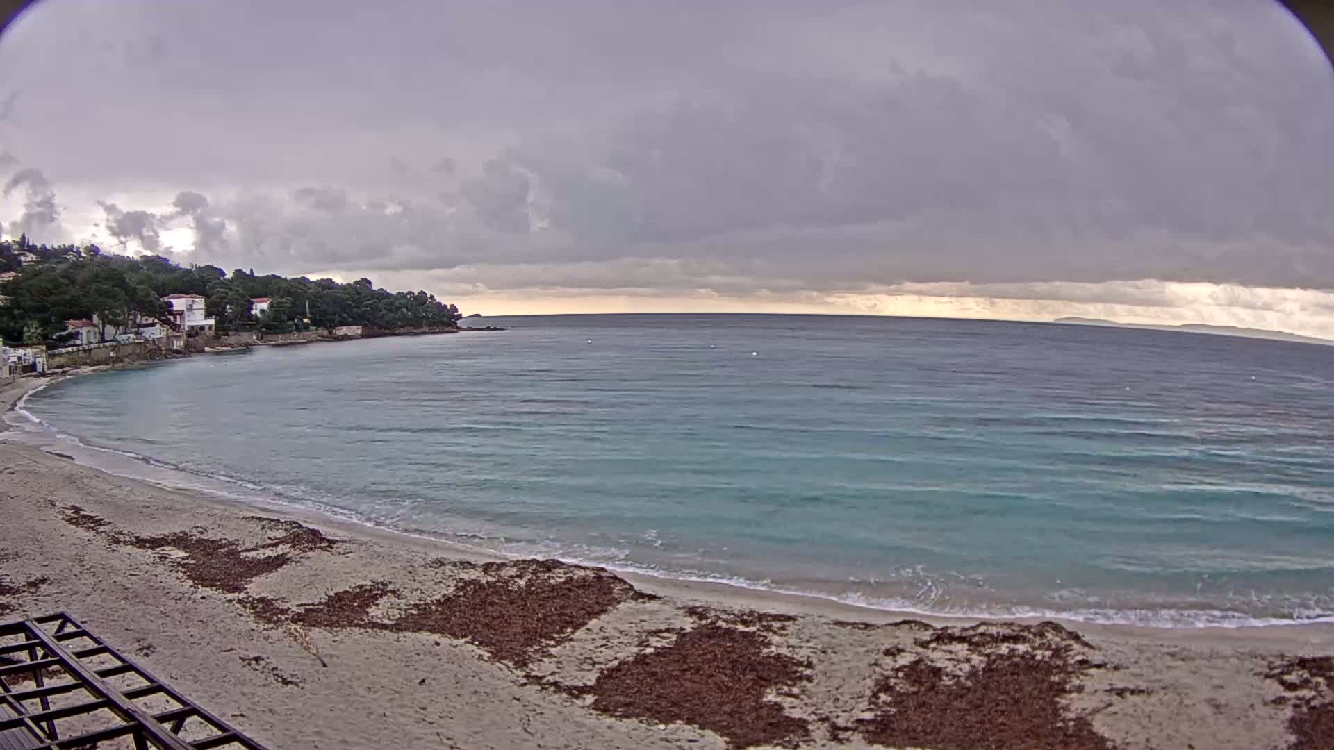 A sandy beach with seaweed washes up to a calm, teal sea under a cloudy, overcast sky.