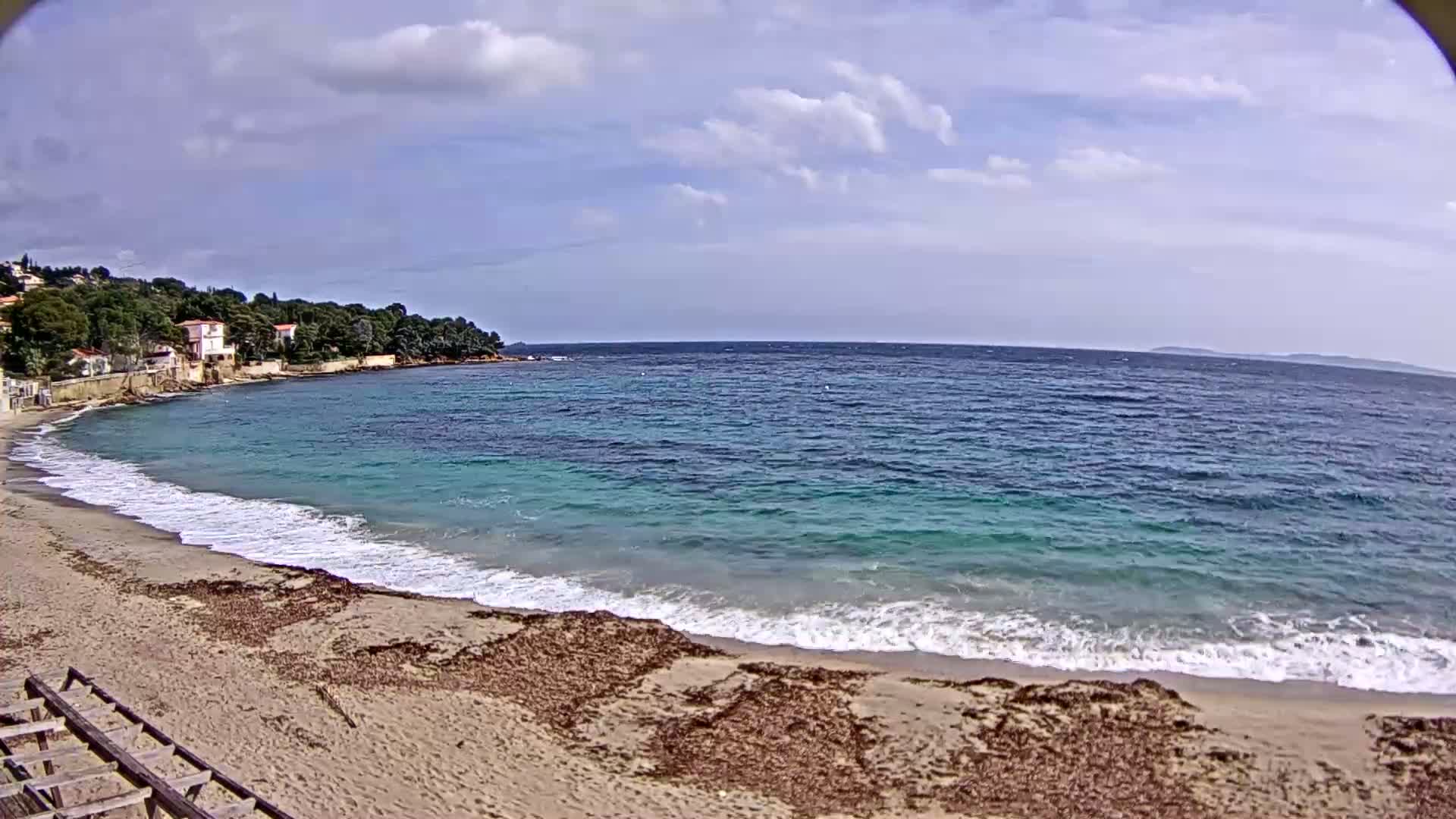 A sandy beach with small waves laps the shore under a partly cloudy sky, with houses and trees visible on a nearby hill.