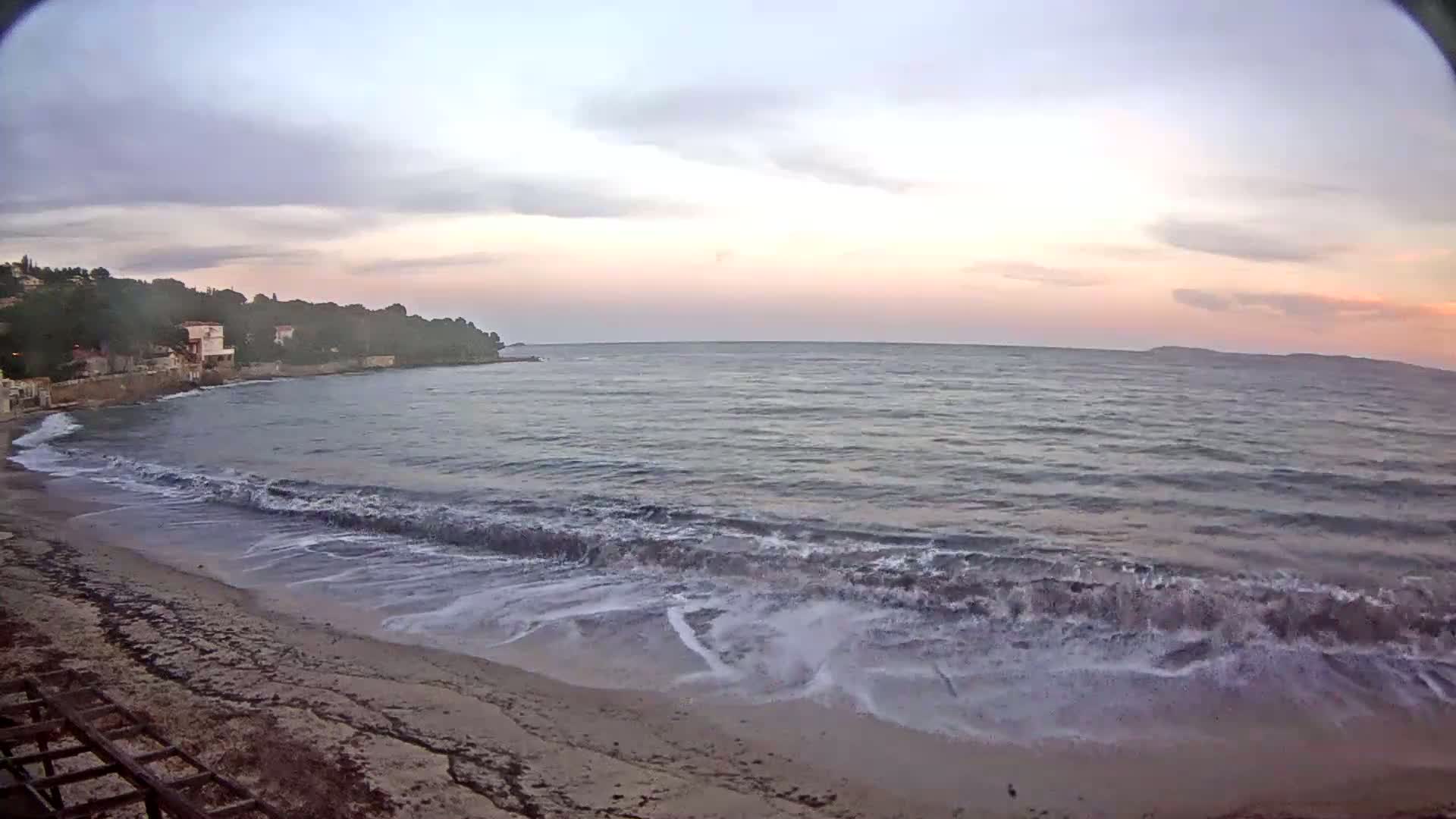 Small waves lap a sandy beach under a pale, sunset sky near a line of houses and trees.