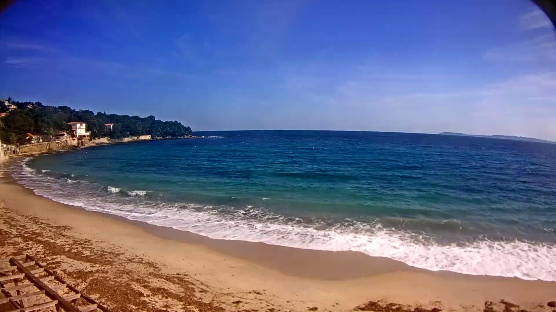 A sandy beach with small waves laps the shore under a clear blue sky, backed by a line of houses and trees.